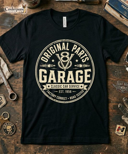 Workbench with a black graphic tee featuring original Cinderpoint Studios artwork for Original Parts Garage, a one-color classic car service badge design with bold lettering, V8 emblem, spark plugs, Classic Car Service banner, Factory Correct text, and Road Tested details.
