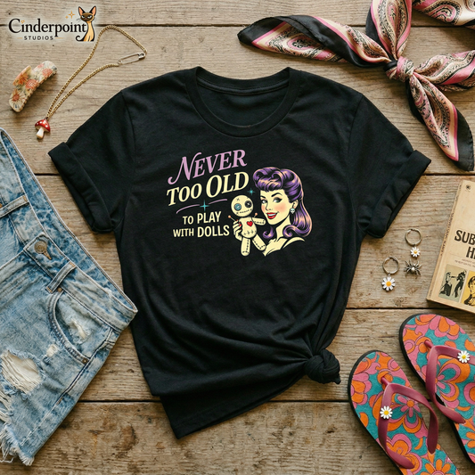 Black retro graphic t-shirt featuring the phrase Never Too Old to Play with Dolls beside a vintage-inspired woman with purple hair holding a stitched voodoo doll.