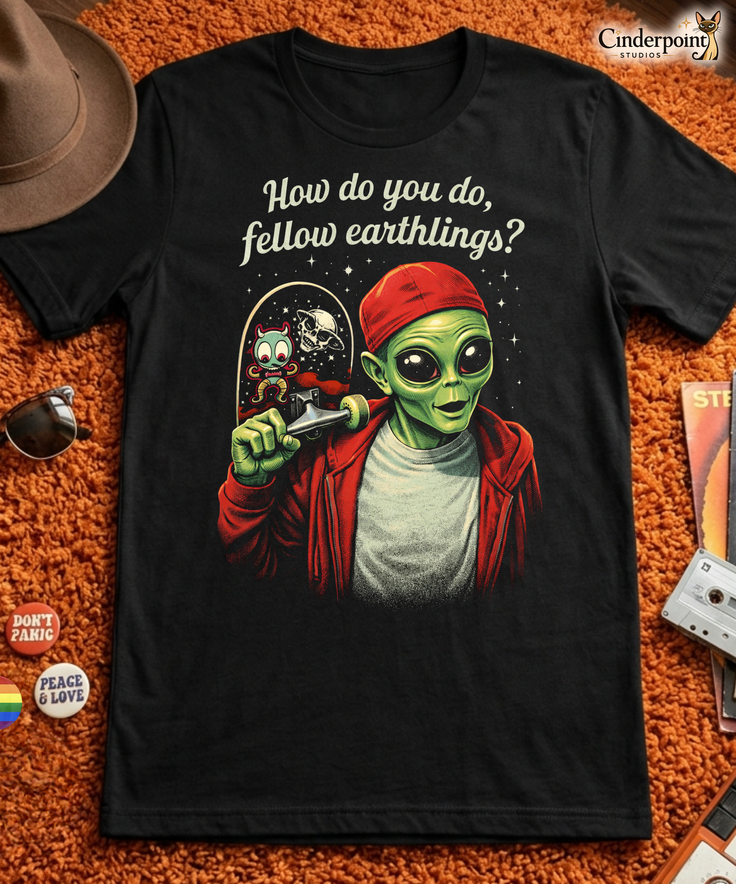 A retro-style graphic on a black t-shirt featuring a green alien on a skateboard, wearing a red hat and holding a skateboard, with the text "FELLOW EARTHLINGS.
