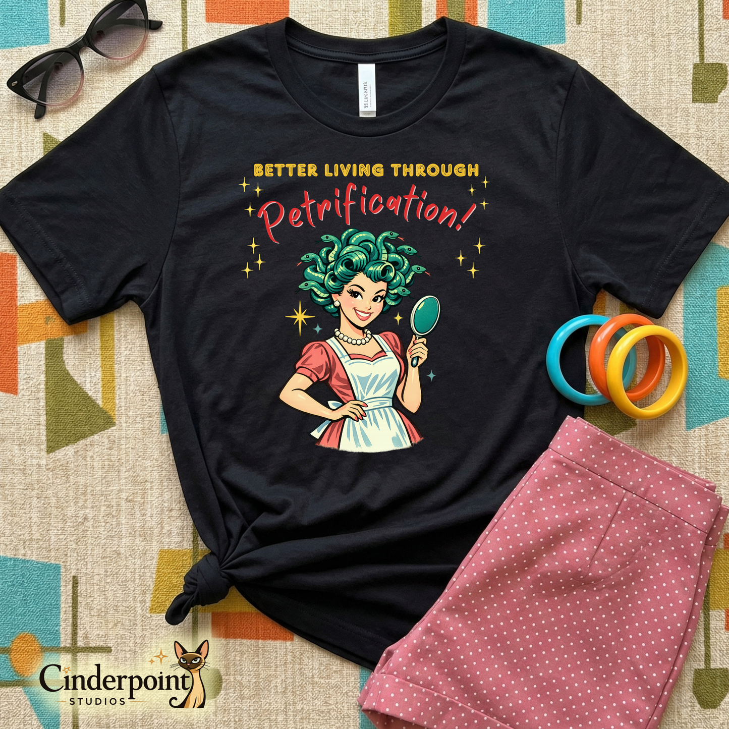 Better Living Through Petrification unisex tee — vintage 1950s retro Medusa housewife with styled snake hair holding a hand mirror and smiling confidently in pearls and apron, gold and red typography on black Bella Canvas 3001 by Cinderpoint Studios