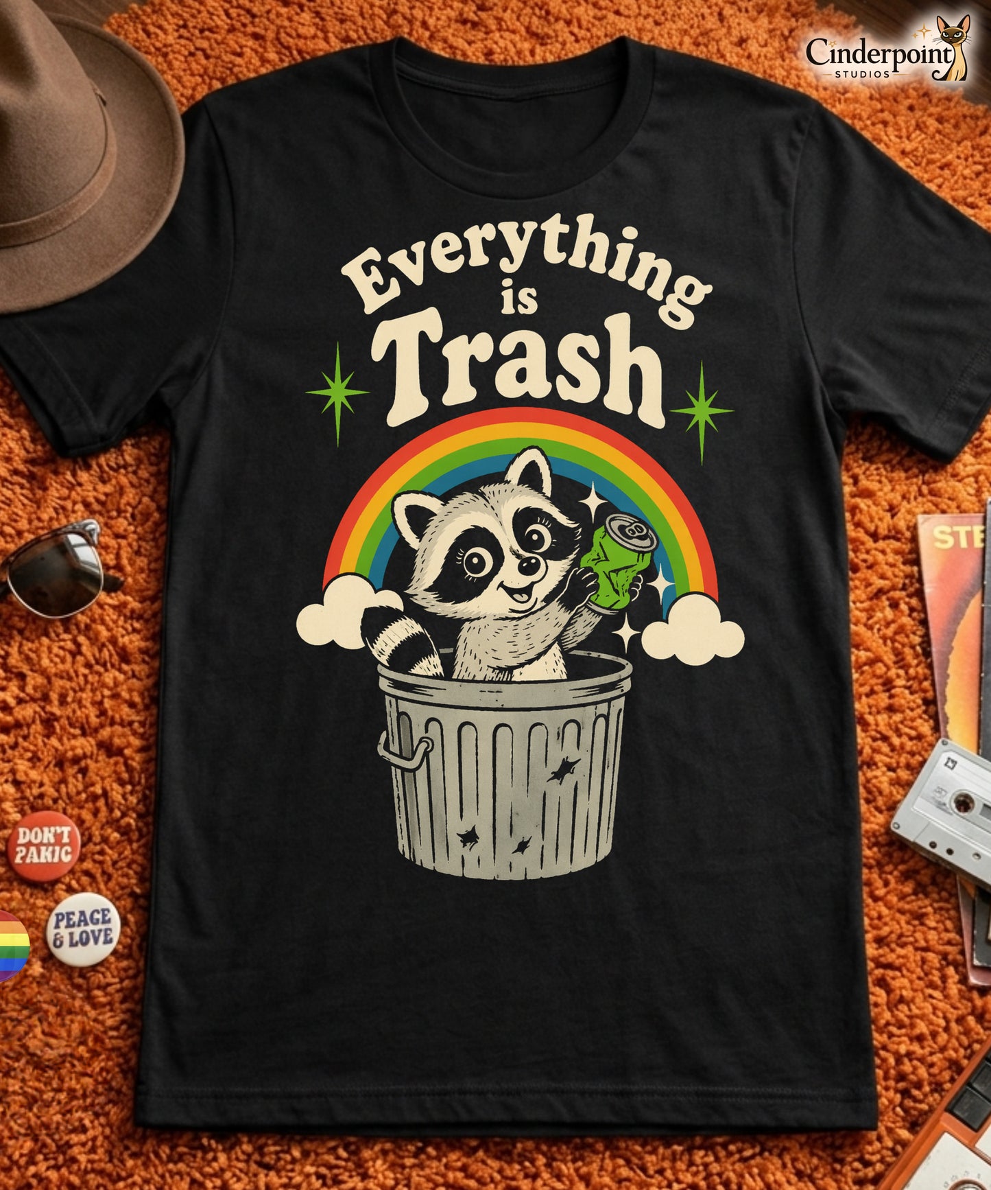 A retro 1970s-style graphic t-shirt featuring a cute raccoon sitting under a vibrant rainbow with the text "Everything Is Trash" in vintage-inspired lettering