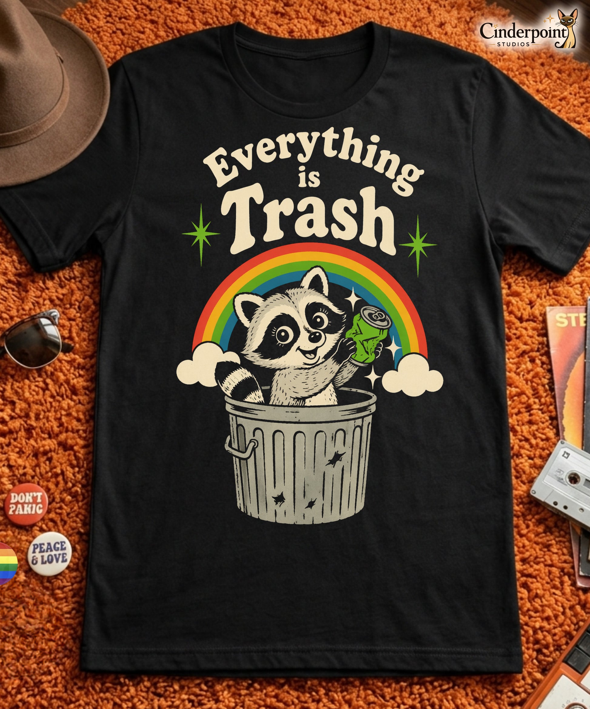 A retro 1970s-style graphic t-shirt featuring a cute raccoon sitting under a vibrant rainbow with the text "Everything Is Trash" in vintage-inspired lettering