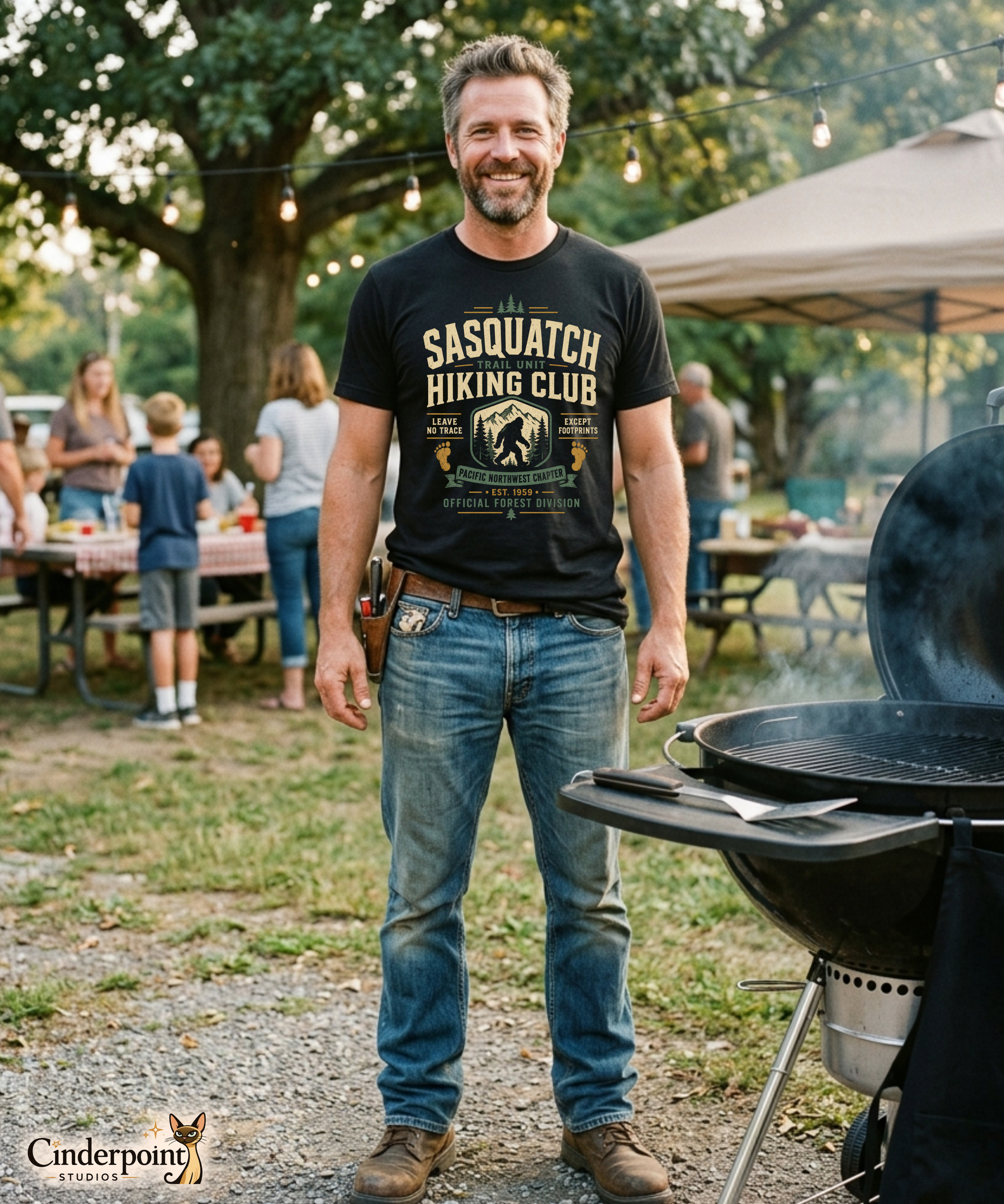 Male model wearing a black graphic tee with the Sasquatch Hiking Club design by Cinderpoint Studios, featuring retro outdoor-club lettering, Bigfoot-inspired artwork, pine trees, and Pacific Northwest trail details.