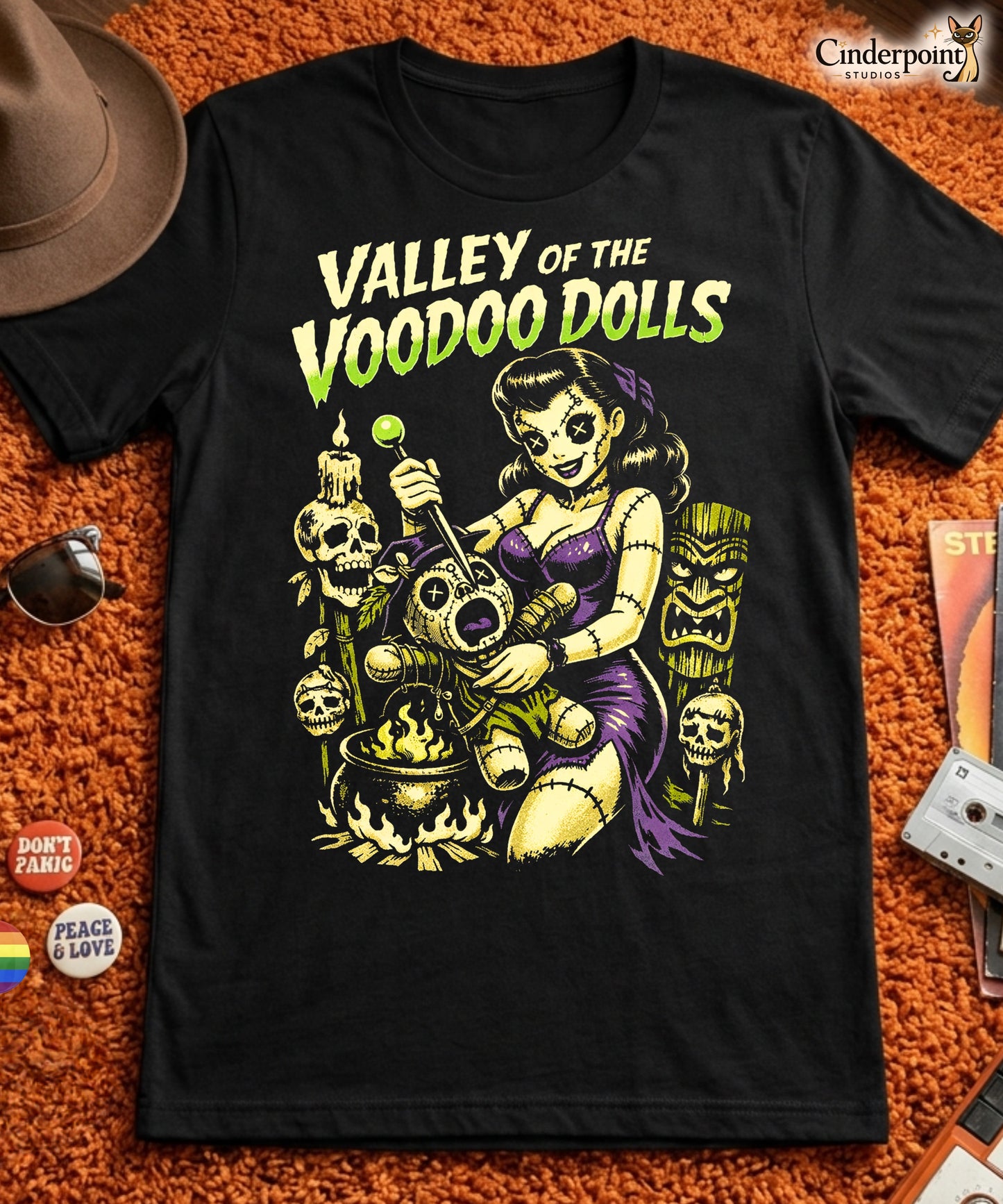 A retro-style graphic on a black t-shirt featuring a stylized pinup girl voodoo doll turning the tables on a medicine man in a vibrant, gritty Tiki-horror aesthetic