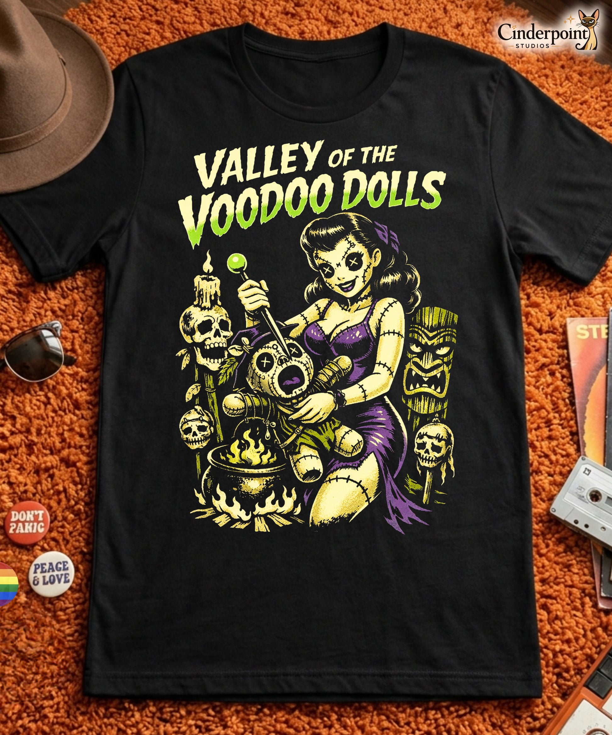 A retro-style graphic on a black t-shirt featuring a stylized pinup girl voodoo doll turning the tables on a medicine man in a vibrant, gritty Tiki-horror aesthetic