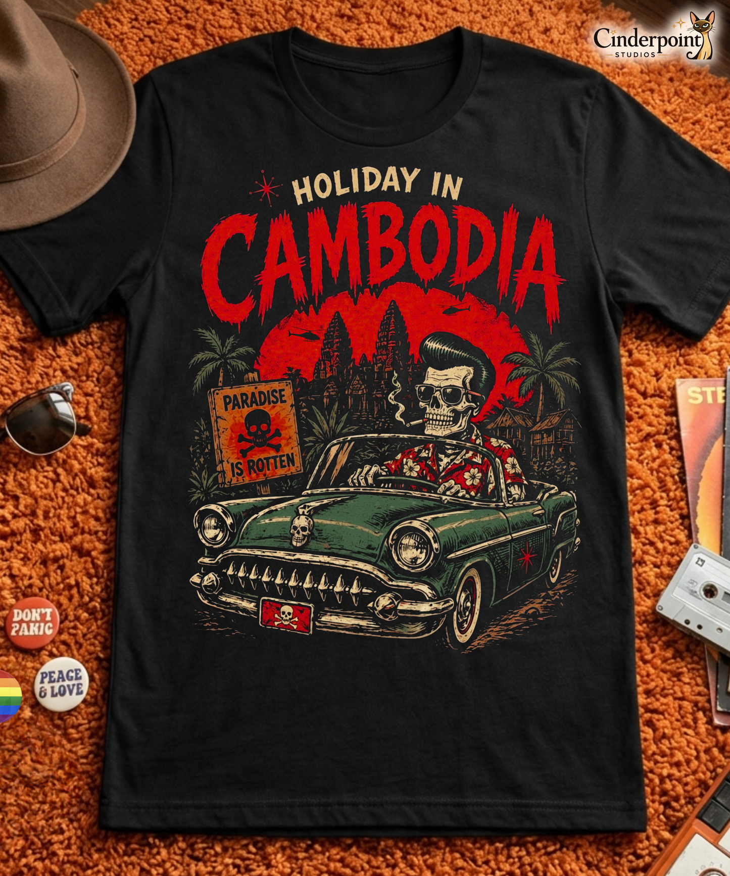 A detailed lowbrow graphic on a black t-shirt featuring a skeleton with a pompadour driving a 1955 Chevy convertible. The background shows Angkor Wat under a red sun with helicopters, and a sign that reads 'Paradise is Rotten'.