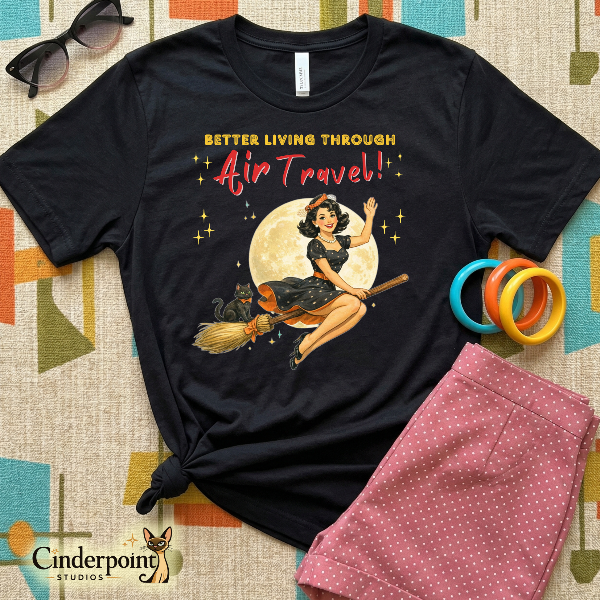 Black t-shirt with a graphic design of a woman on a broomstick, text 'Better Living Through Air Travel', sunglasses, colorful rings, and pink polka dot shorts on a patterned surface.