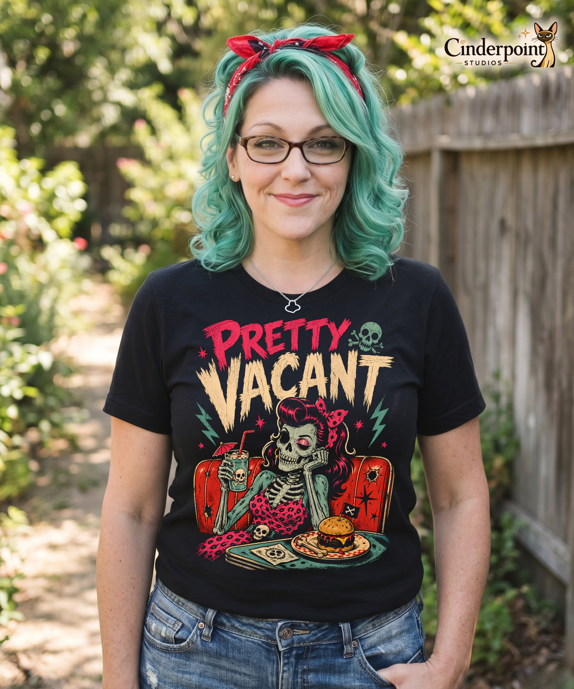 A retro-style graphic on a black t-shirt of a female skeleton pinup with pink hair in a diner booth, holding a skull-themed milkshake next to a burger with a crossbones flag.