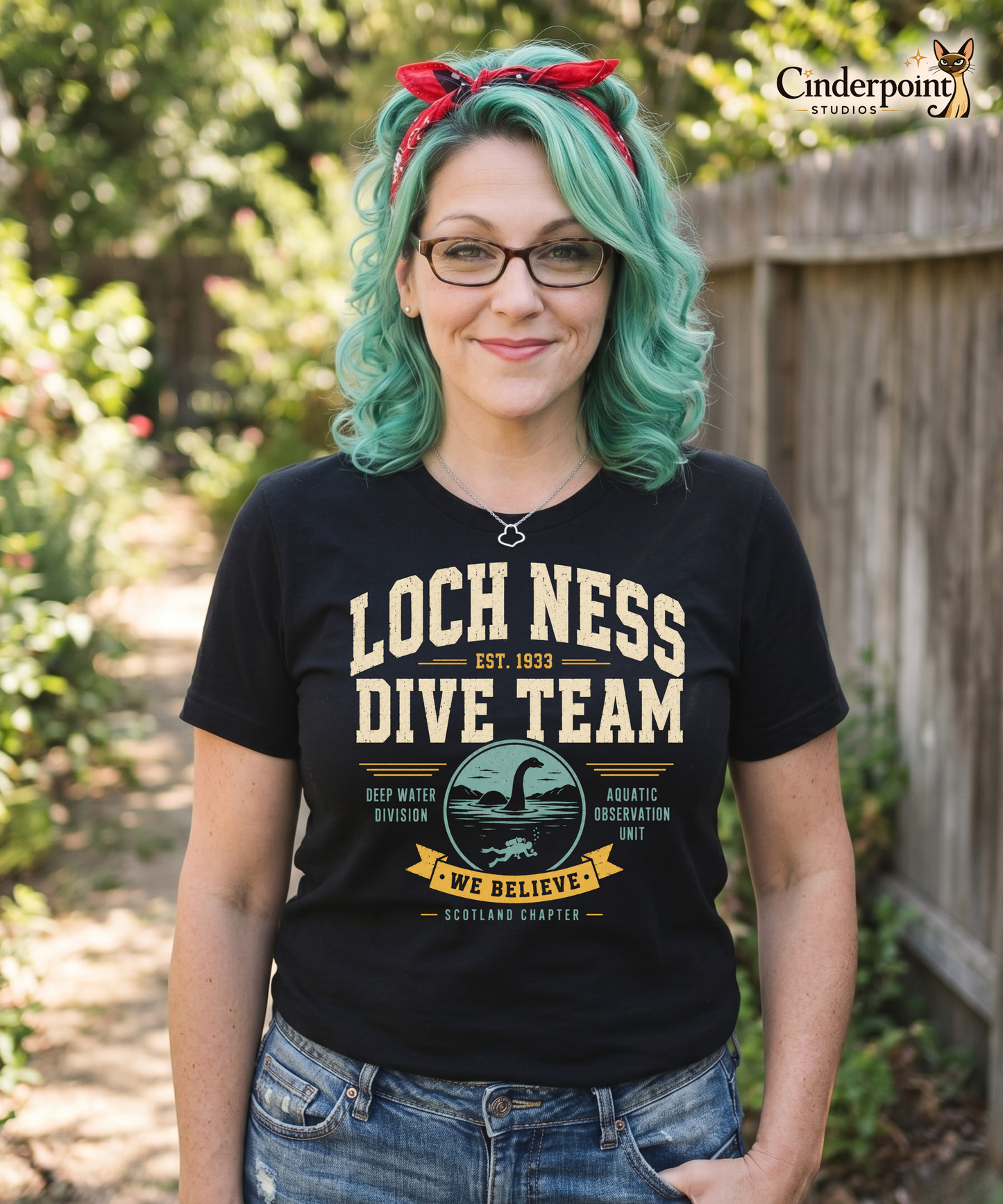 Woman wearing a Black graphic tee featuring original Cinderpoint Studios artwork for Loch Ness Dive Team, a retro cryptid design with bold varsity lettering, a Nessie-style lake monster, diver, water scene, and text including Deep Water Division, Aquatic Observation Unit, We Believe, and Scotland Chapter.