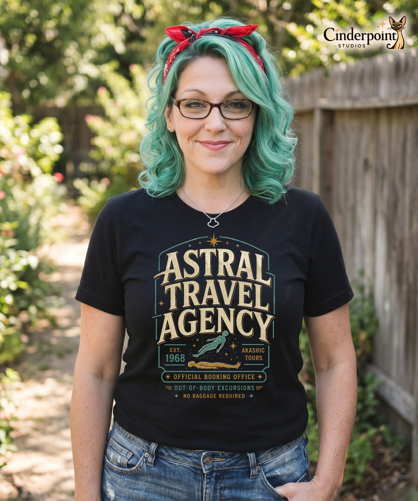 Woman wearing a black graphic tee featuring original Cinderpoint Studios artwork for Astral Travel Agency, a retro occult travel design with large vintage lettering, astral projection imagery, stars, and text including Official Booking Office, Akashic Tours, and Out-of-Body Excursions.