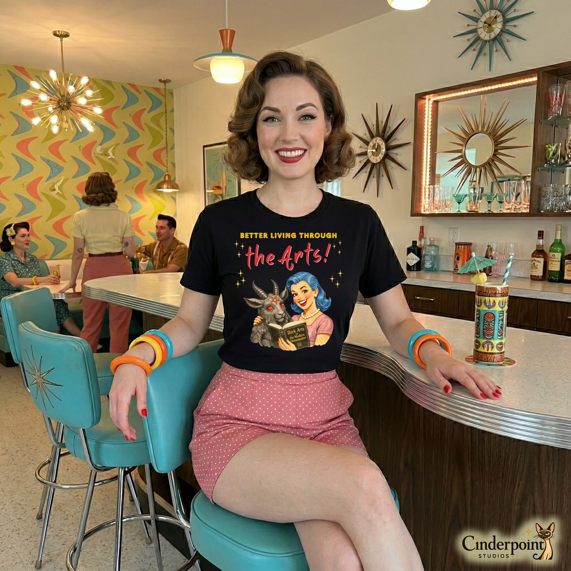 A woman sitting at a counter in a retro-style diner wearing a t-shirt with an artistic design. Retro watercolor illustration of a woman with blue hair and pearls reading a "Dark Arts" book next to a goat with a pentagram on its forehead on a black t-shirt.