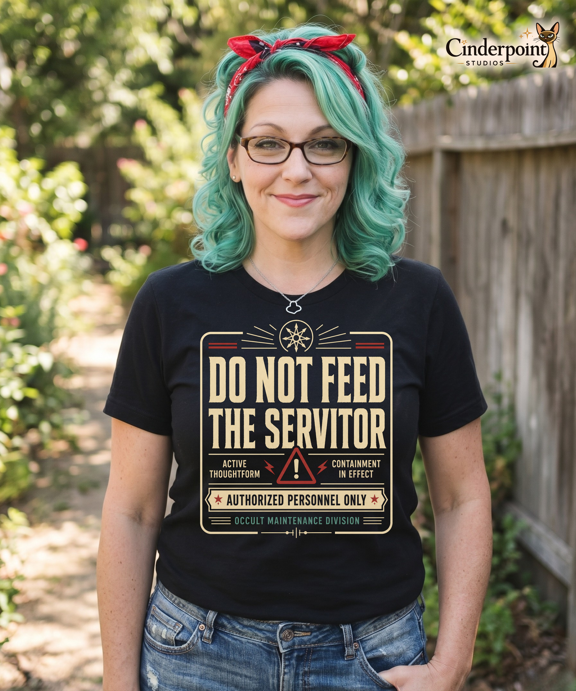 Auntie Mel is wearing a black graphic tee featuring original Cinderpoint Studios artwork for Do Not Feed the Servitor, a retro occult warning-sign design with bold vintage lettering, a chaos star symbol, a hazard icon, and text including Active Thoughtform, Containment in Effect, Authorized Personnel Only, and Occult Maintenance Division.
