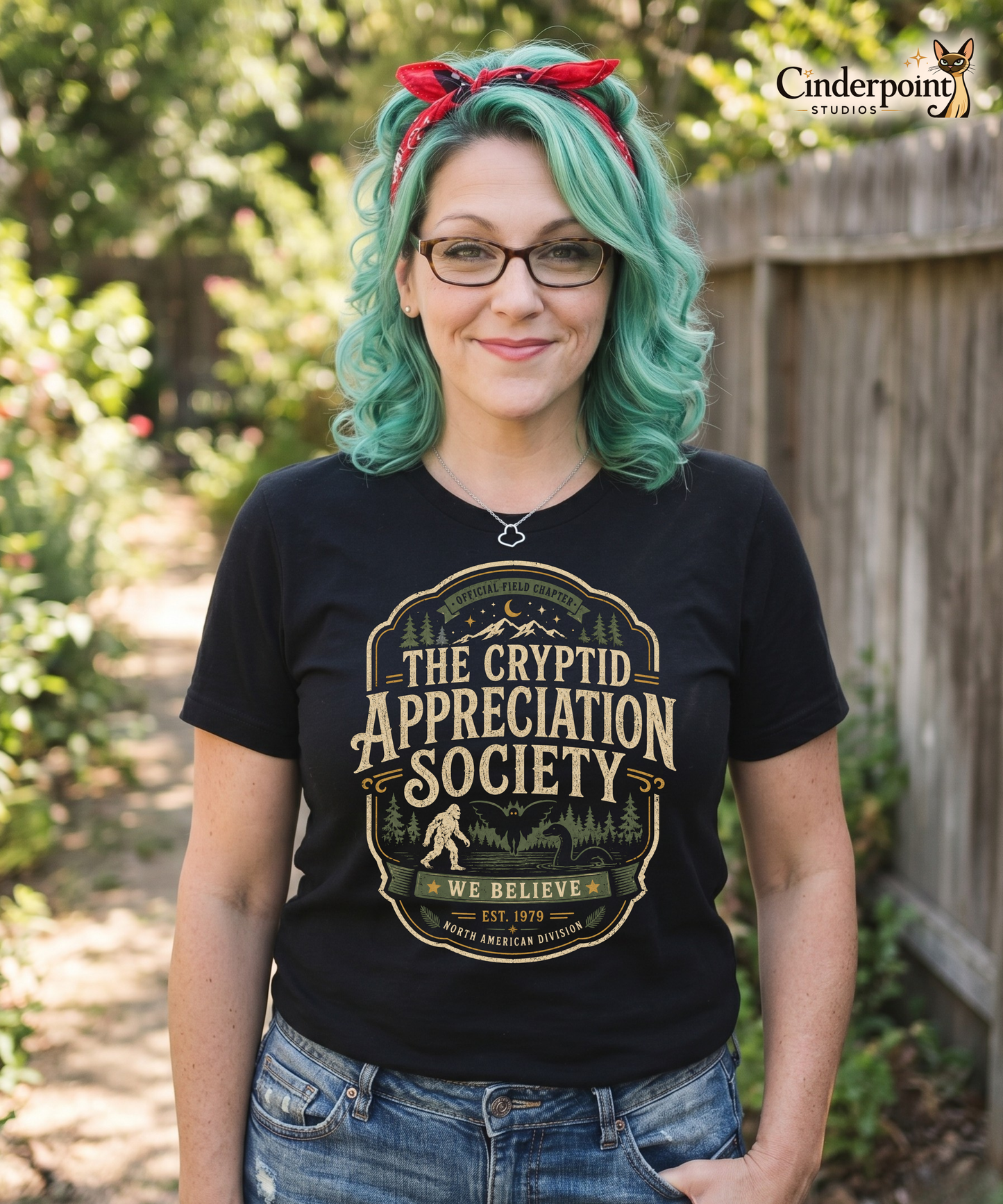 Female model wearing a black graphic tee with The Cryptid Appreciation Society artwork by Cinderpoint Studios, styled as a boutique retro paranormal and cryptid fan shirt with Sasquatch, Mothman, Nessie-style creature, and forest badge artwork.