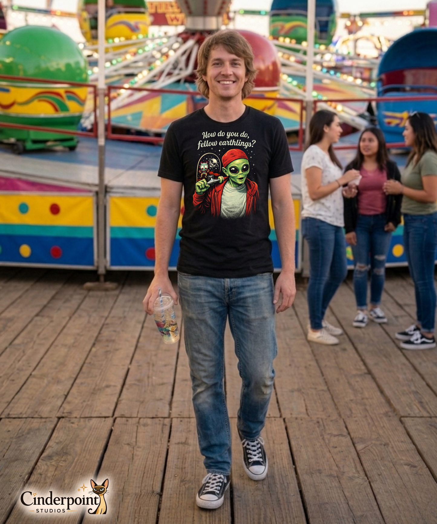 A man at a carnival wearing a retro-style graphic on a black t-shirt featuring a green alien on a skateboard, wearing a red hat and holding a skateboard, with the text "FELLOW EARTHLINGS.