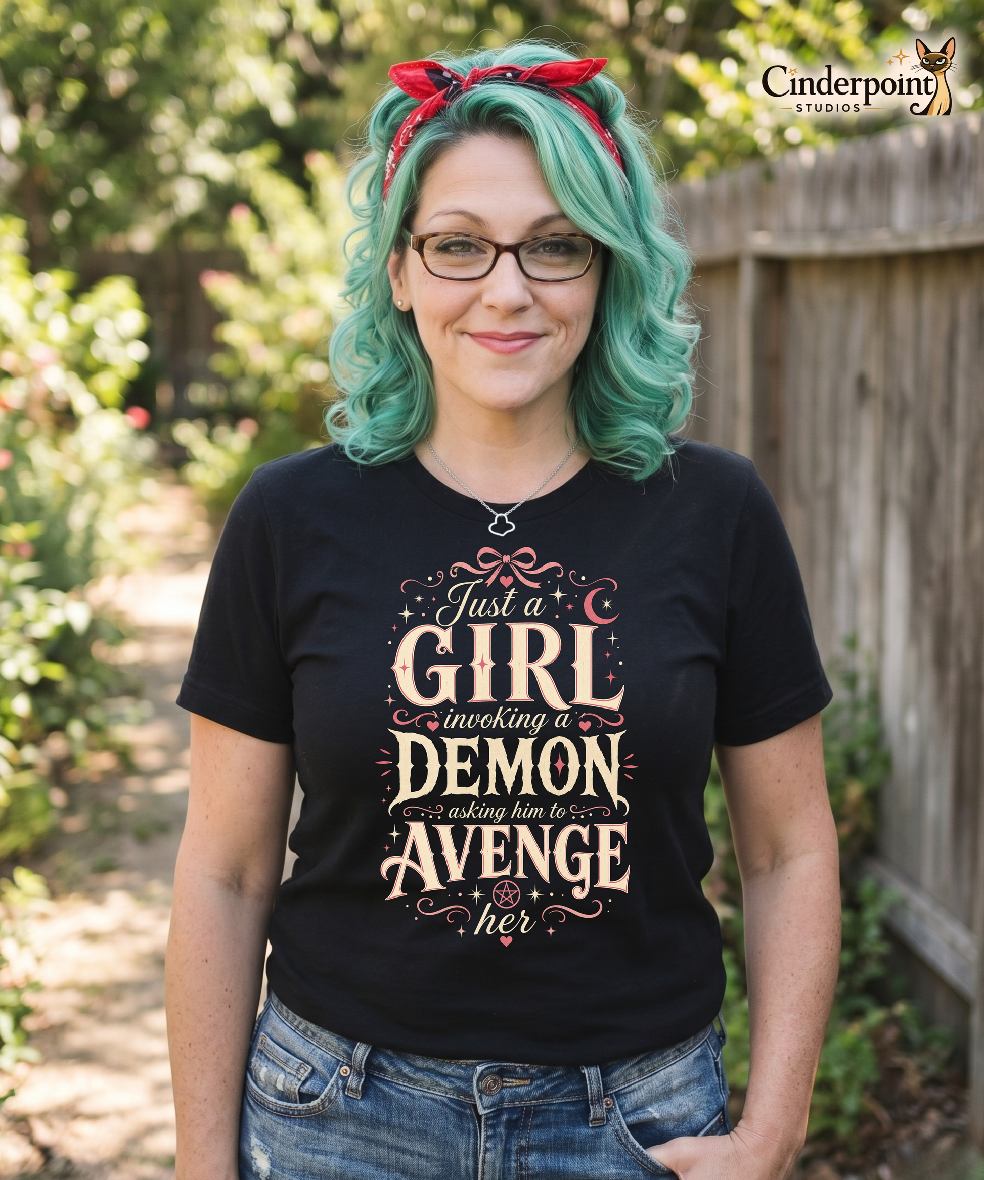 Auntie Mel, the designer, wearing a black graphic tee featuring original Cinderpoint Studios artwork for Just a Girl Invoking a Demon, a feminine occult humor design with bold vintage lettering, pink bow accents, crescent moon, stars, hearts, and a small pentagram.
