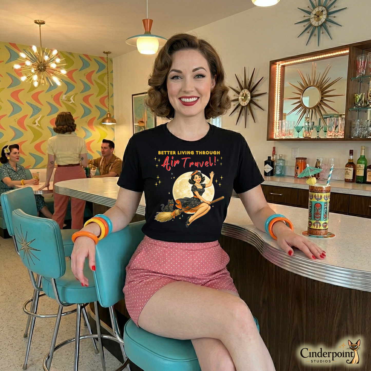 Woman sitting at a counter in a retro-style diner wearing a t-shirt with vintage design. Better Living Through Air Travel unisex tee — vintage retro pinup witch in black polka dot dress and stewardess hat riding a broom in front of a full moon with a dapper black cat familiar in orange bow tie, gold and red typography on black Bella Canvas 3001 by Cinderpoint Studios