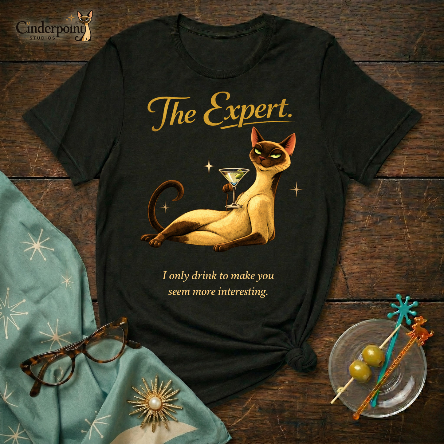 A black Bella+Canvas 3001 t-shirt featuring "The Expert" design on a dark wood grain table. The design shows a Siamese cat lounging with a martini and the text "The Expert" and "I only drink to make you seem more interesting." Accessories include vintage cat-eye glasses and a cocktail garnish dish. Cinderpoint Studios logo in the corner.