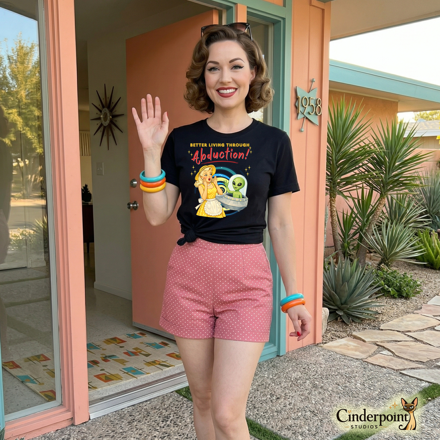 Woman wearing a black t-shirt with a graphic design and pink shorts, standing in front of a house entrance. Retro watercolor illustration of a 1950s woman in a yellow polka dot dress waving at a friendly green alien in a UFO on a black t-shirt.