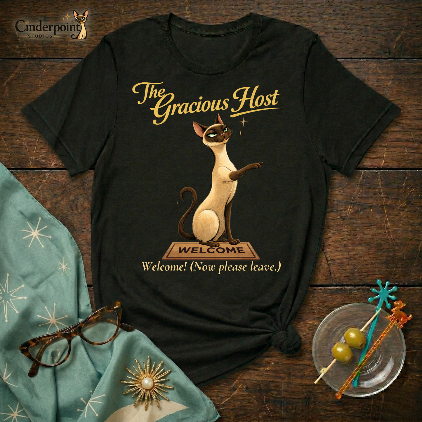 A black Bella+Canvas 3001 t-shirt featuring "The Gracious Host" design. The artwork shows Lyle the Siamese cat sitting regally behind a wooden "Welcome" mat with a smug, knowing expression. The text reads "The Gracious Host" and "Welcome! (Now please leave.)" placed on a dark wood grain surface with vintage accessories.