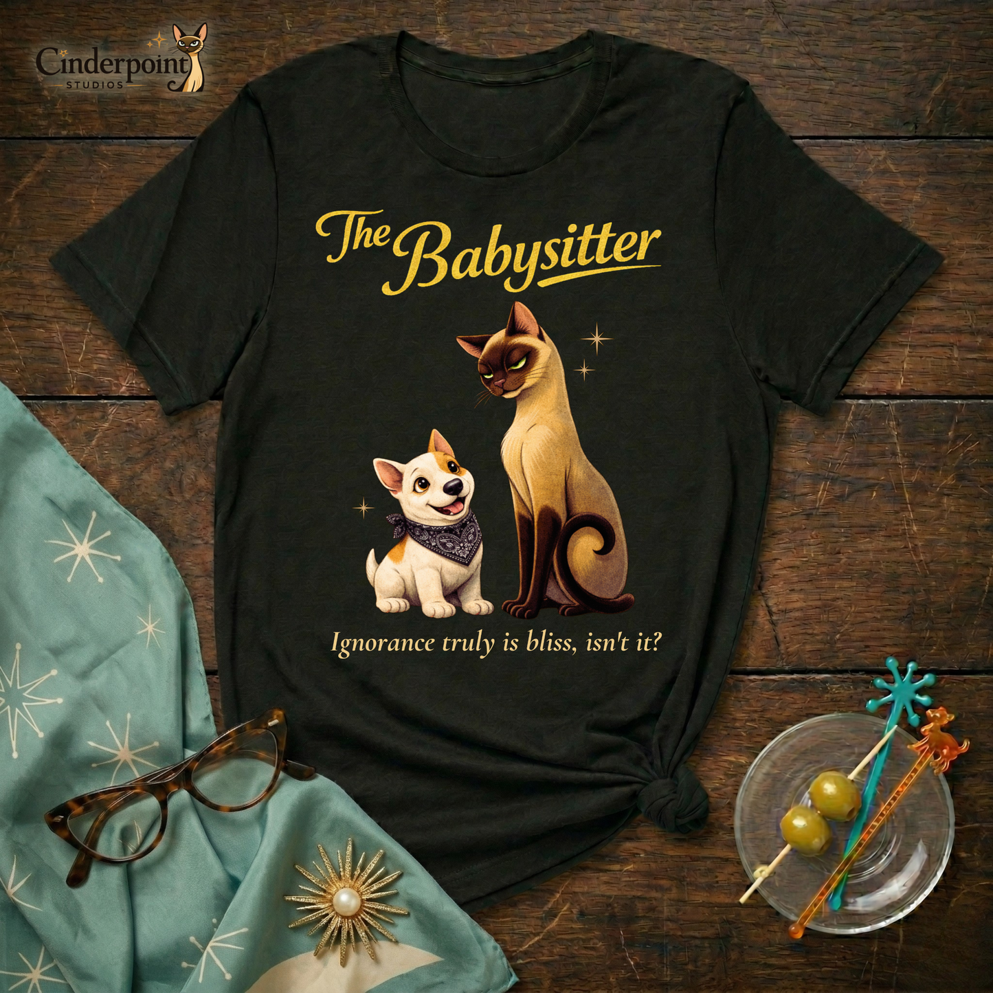 A black Bella+Canvas 3001 t-shirt featuring "The Babysitter" design on a dark wood grain table. The design shows a judgmental Siamese cat looking down at a happy Bull Terrier puppy with the text "The Babysitter" and "Ignorance truly is bliss, isn't it?" Accessories include vintage glasses and a starburst scarf.