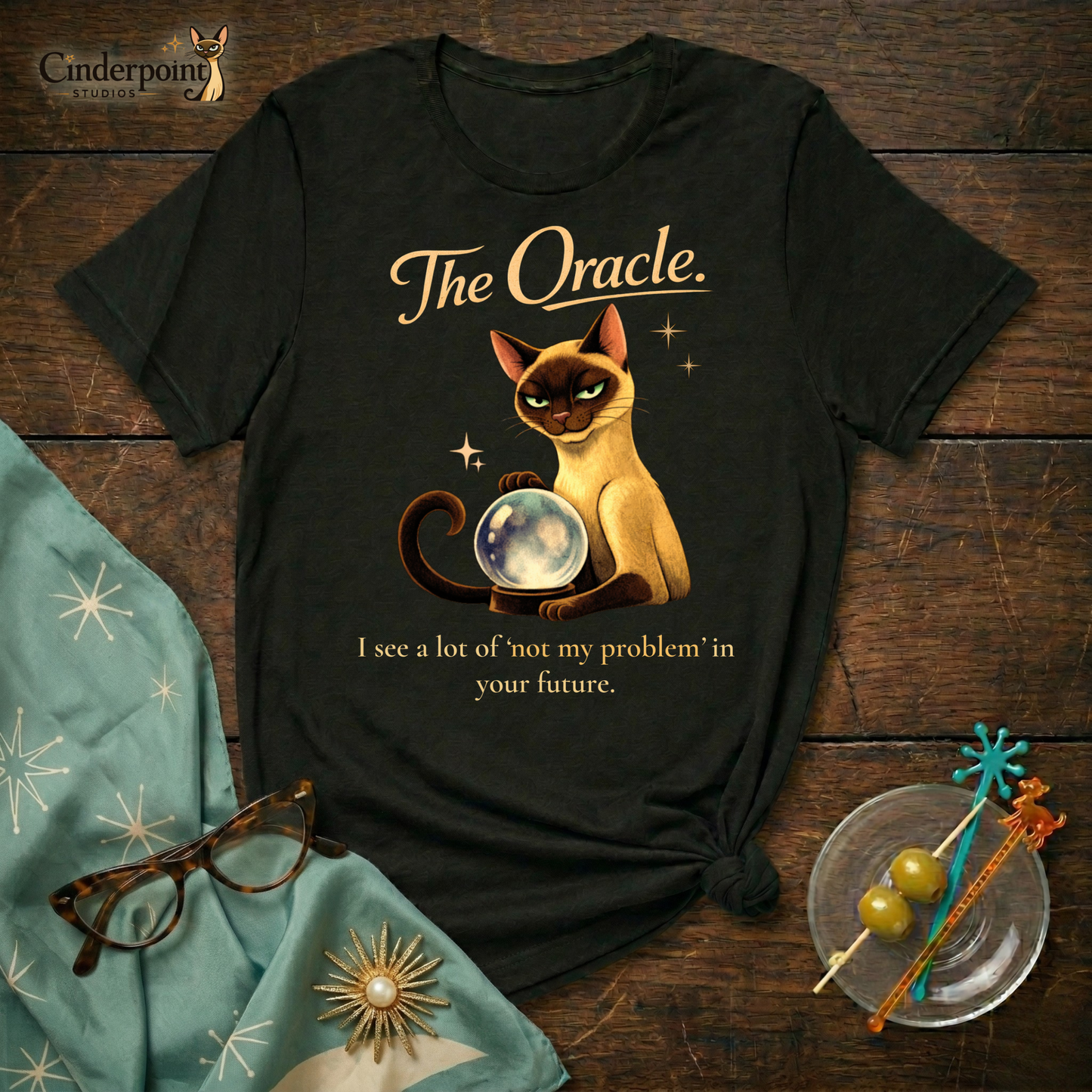 A black Bella+Canvas 3001 t-shirt featuring "The Oracle" design on a dark wood grain table. The design shows a Siamese cat surrounded by atomic stars and the text "The Oracle" and "I see your problem. I also see that it’s not mine." Accessories include vintage cat-eye glasses and a starburst scarf. Cinderpoint Studios logo in the corner.