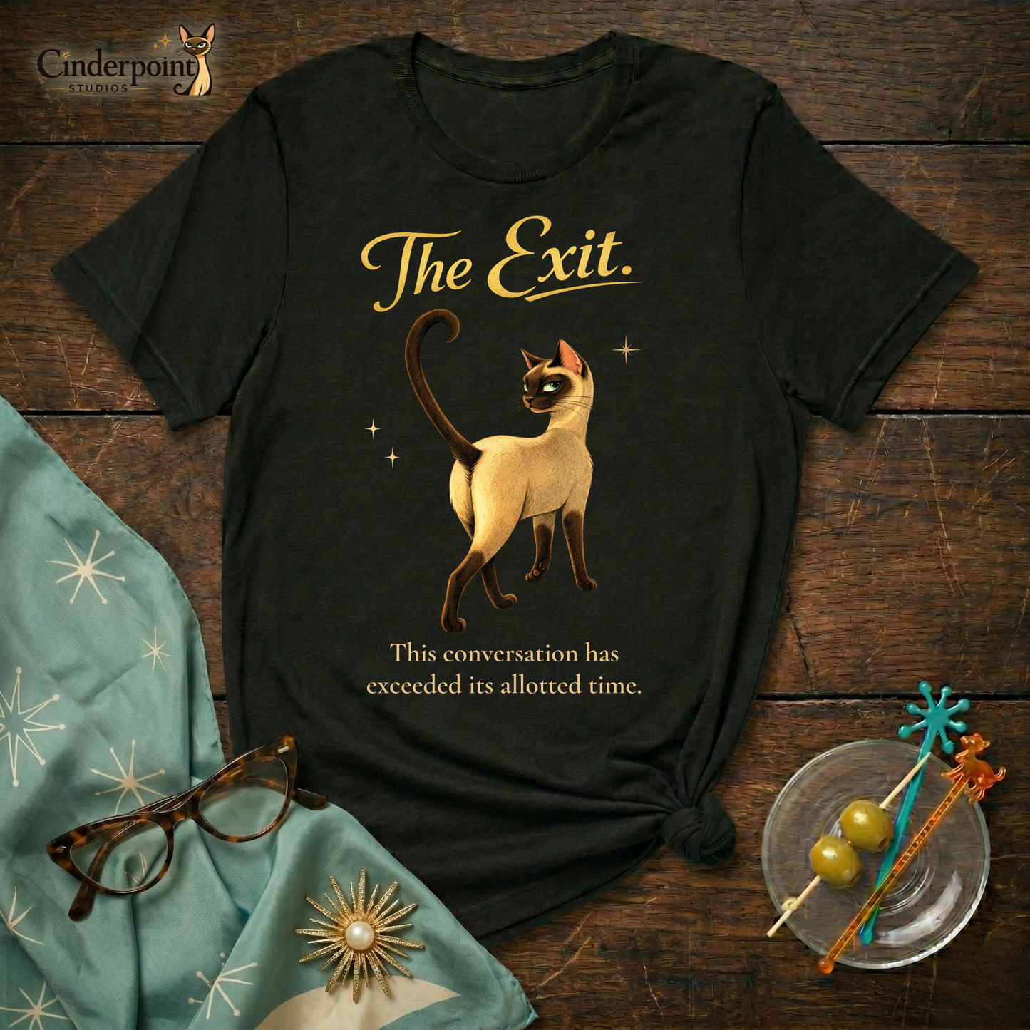 A black Bella+Canvas 3001 t-shirt featuring "The Exit" design on a dark wood grain table. The design shows a Siamese cat walking away with the text "The Exit" and "This conversation has exceeded its allotted time." Accessories include turquoise cat-eye glasses, a matching starburst scarf, and a glass dish with olives. Cinderpoint Studios logo in the corner.