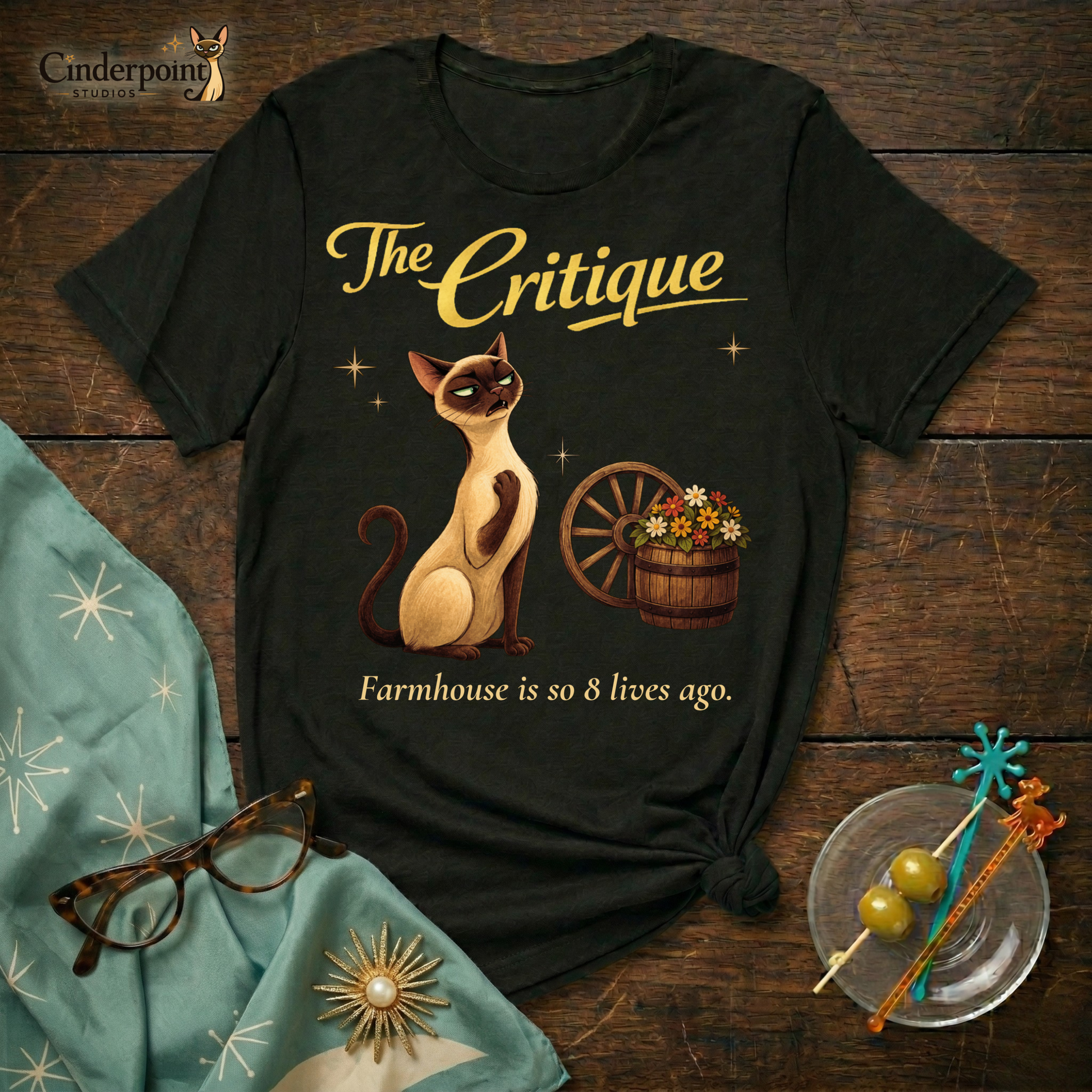 A black Bella+Canvas 3001 t-shirt featuring "The Critique" design. The artwork shows Lyle the Siamese cat recoiling in horror from a rustic wagon wheel and flower barrel. The text reads "The Critique" and "Farmhouse is so 8 lives ago." Placed on a dark wood grain surface with vintage barware and atomic starburst accessories.