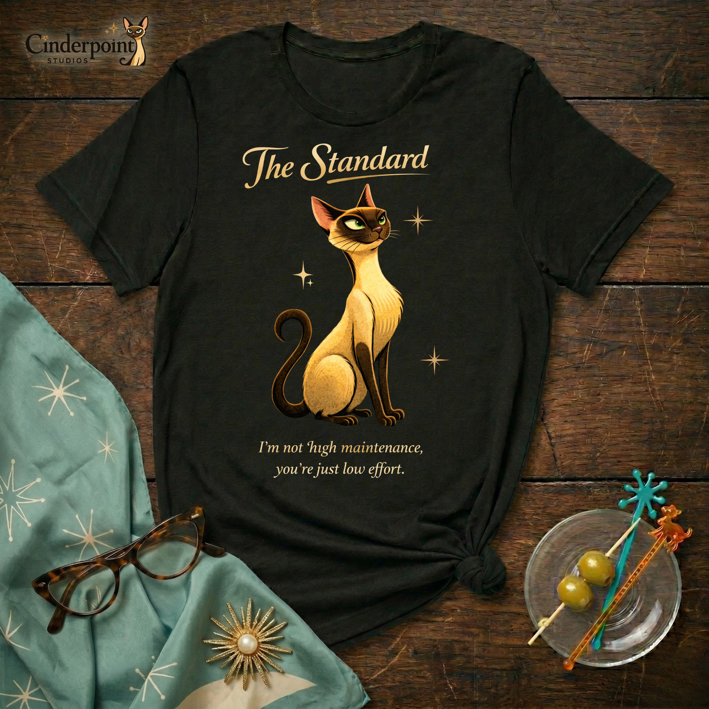 A black Bella+Canvas 3001 t-shirt featuring "The Standard" design, placed on a dark wood grain table. The design shows a stylized Siamese cat, the text "The Standard," and the quote "I’m not high maintenance. You’re just low effort." Next to the shirt are accessories: turquoise cat-eye glasses, a matching turquoise atomic starburst scarf, and a small glass dish with green olives on decorative vintage cocktail picks. The Cinderpoint Studios logo is in the top left.