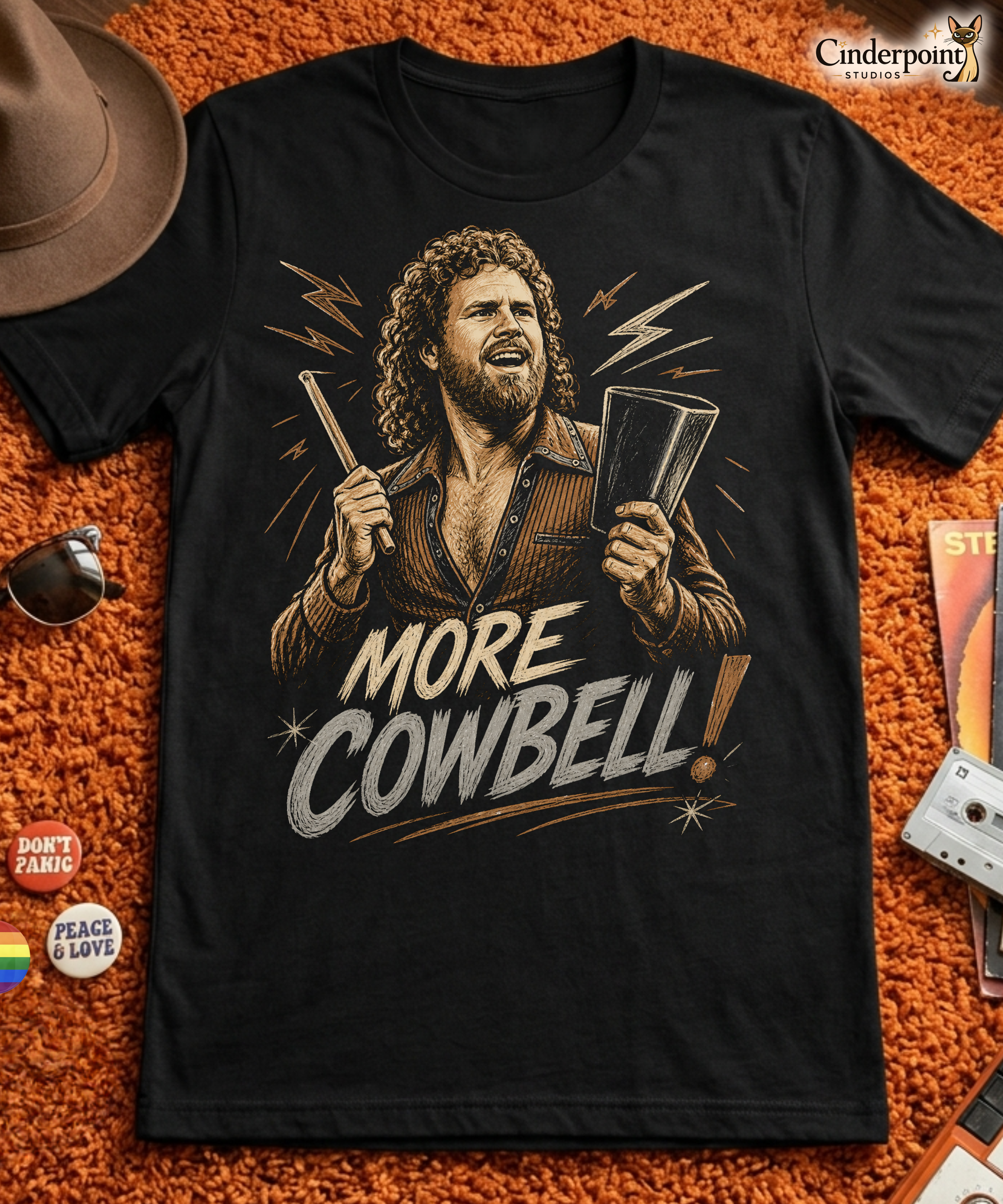 A gritty, woodcut-style illustration on a black t-shirt featuring a bearded man with curly hair and an open-collared 70s shirt holding a cowbell and drumstick, with the text 'MORE COWBELL!' in a distressed font.