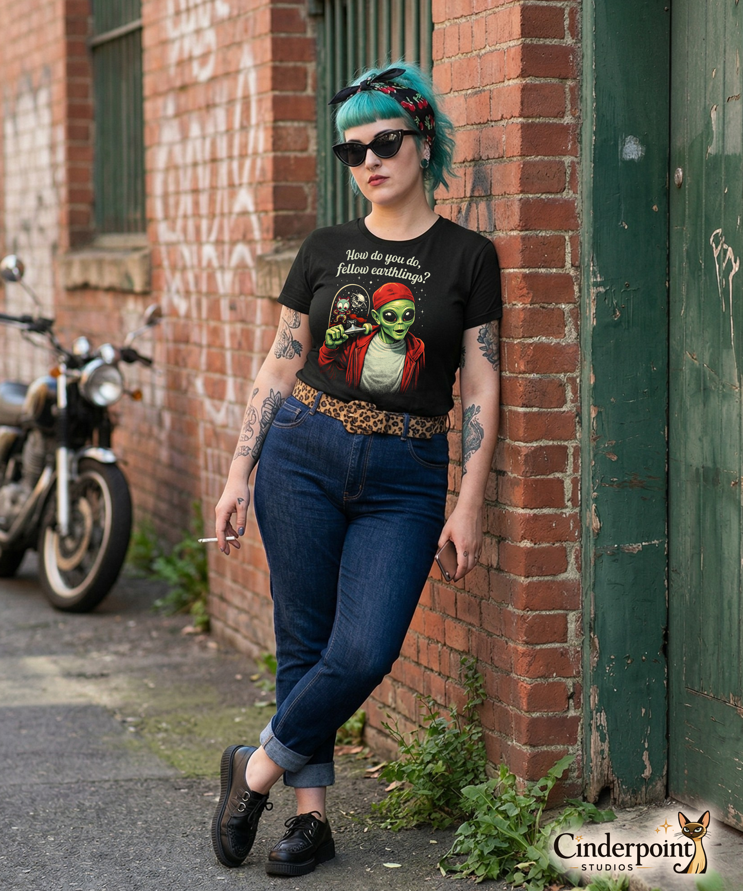 A punk rock girl outdoors wearing a retro-style graphic on a black t-shirt featuring a green alien on a skateboard, wearing a red hat and holding a skateboard, with the text "FELLOW EARTHLINGS.