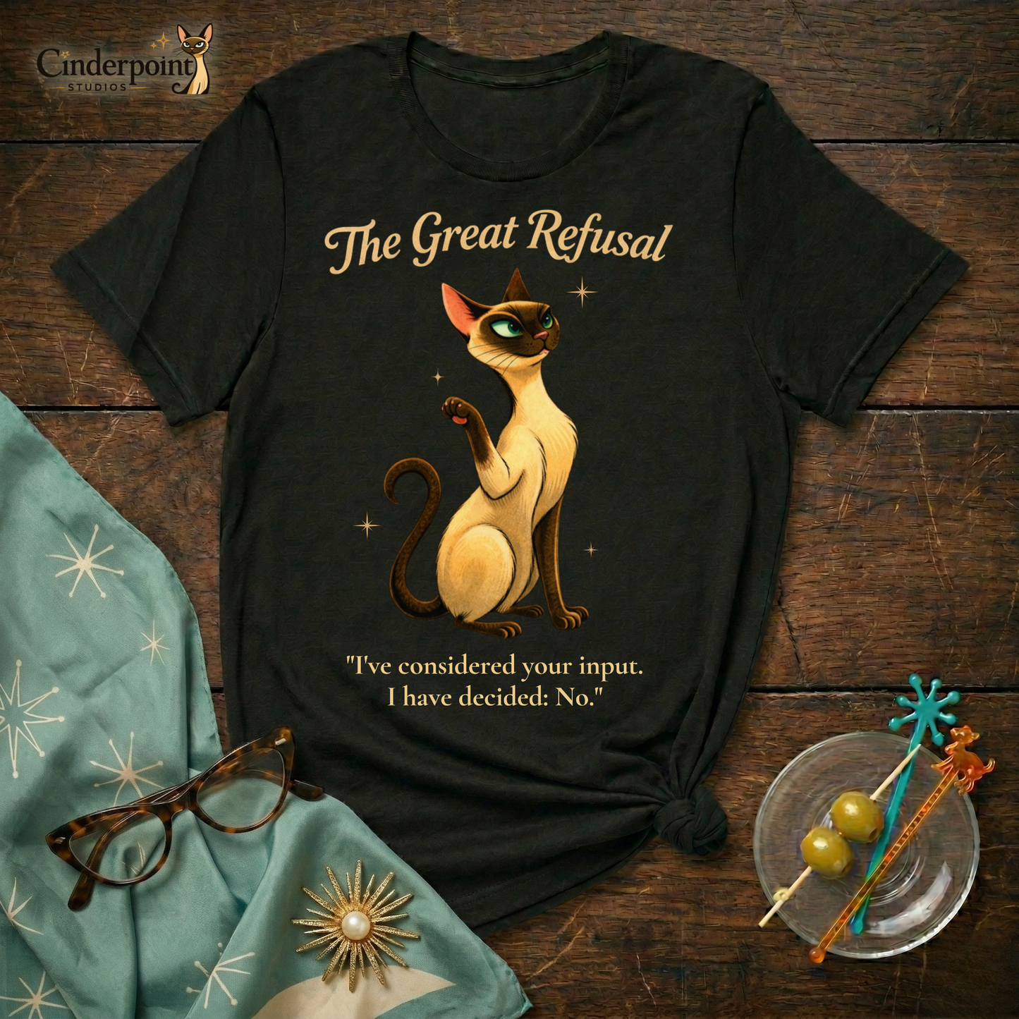 Flat layout on a dark wooden surface. Features a black graphic t-shirt featuring a Siamese cat with his paw up dismissively, and is titled "The Great Refusal." Surrounded by an atomic age scarf with snowflake starbursts, cat-eye glasses, swizzle sticks and two martini olives.
