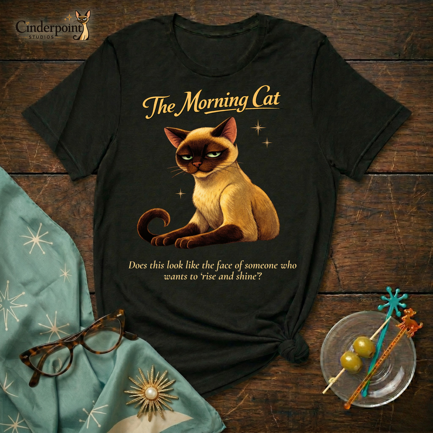 A black Bella+Canvas 3001 t-shirt featuring "The Morning Cat" design on a dark wood grain table. The design shows an exhausted Siamese cat with narrowed eyes and the text "The Morning Cat" and "Does this look like the face of someone who wants to ‘rise and shine’?" Accessories include vintage cat-eye glasses and mid-century modern decor.