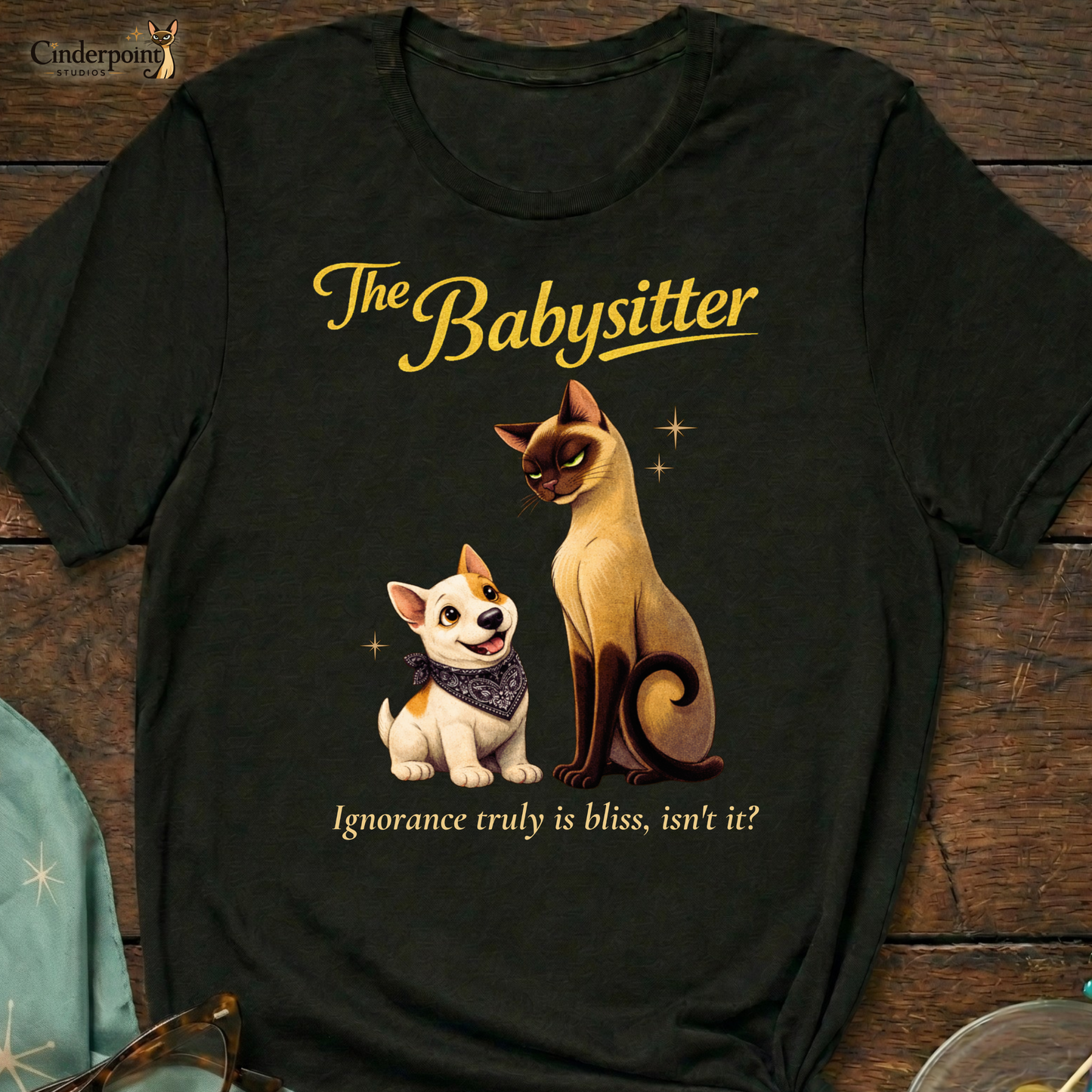A black Bella+Canvas 3001 t-shirt featuring "The Babysitter" design on a dark wood grain table. The design shows a judgmental Siamese cat looking down at a happy Bull Terrier puppy with the text "The Babysitter" and "Ignorance truly is bliss, isn't it?" Accessories include vintage glasses and a starburst scarf.