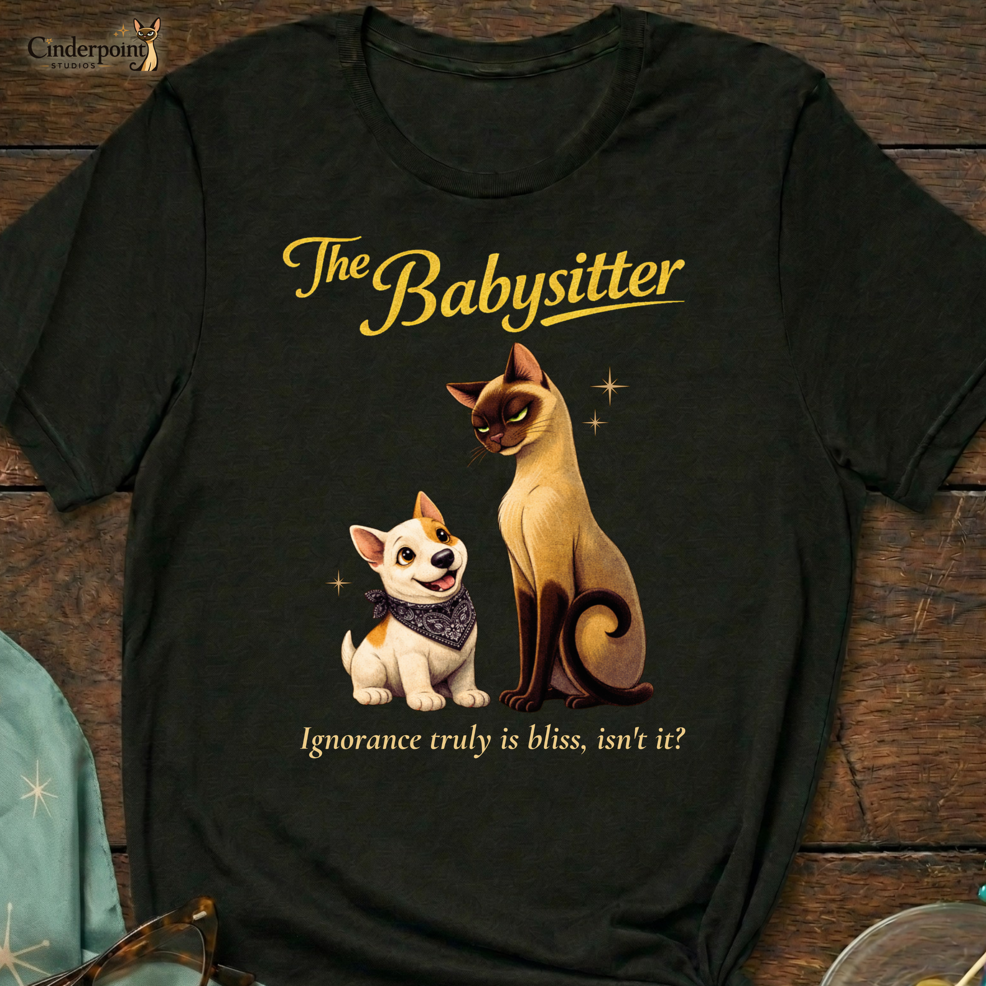 A black Bella+Canvas 3001 t-shirt featuring "The Babysitter" design on a dark wood grain table. The design shows a judgmental Siamese cat looking down at a happy Bull Terrier puppy with the text "The Babysitter" and "Ignorance truly is bliss, isn't it?" Accessories include vintage glasses and a starburst scarf.