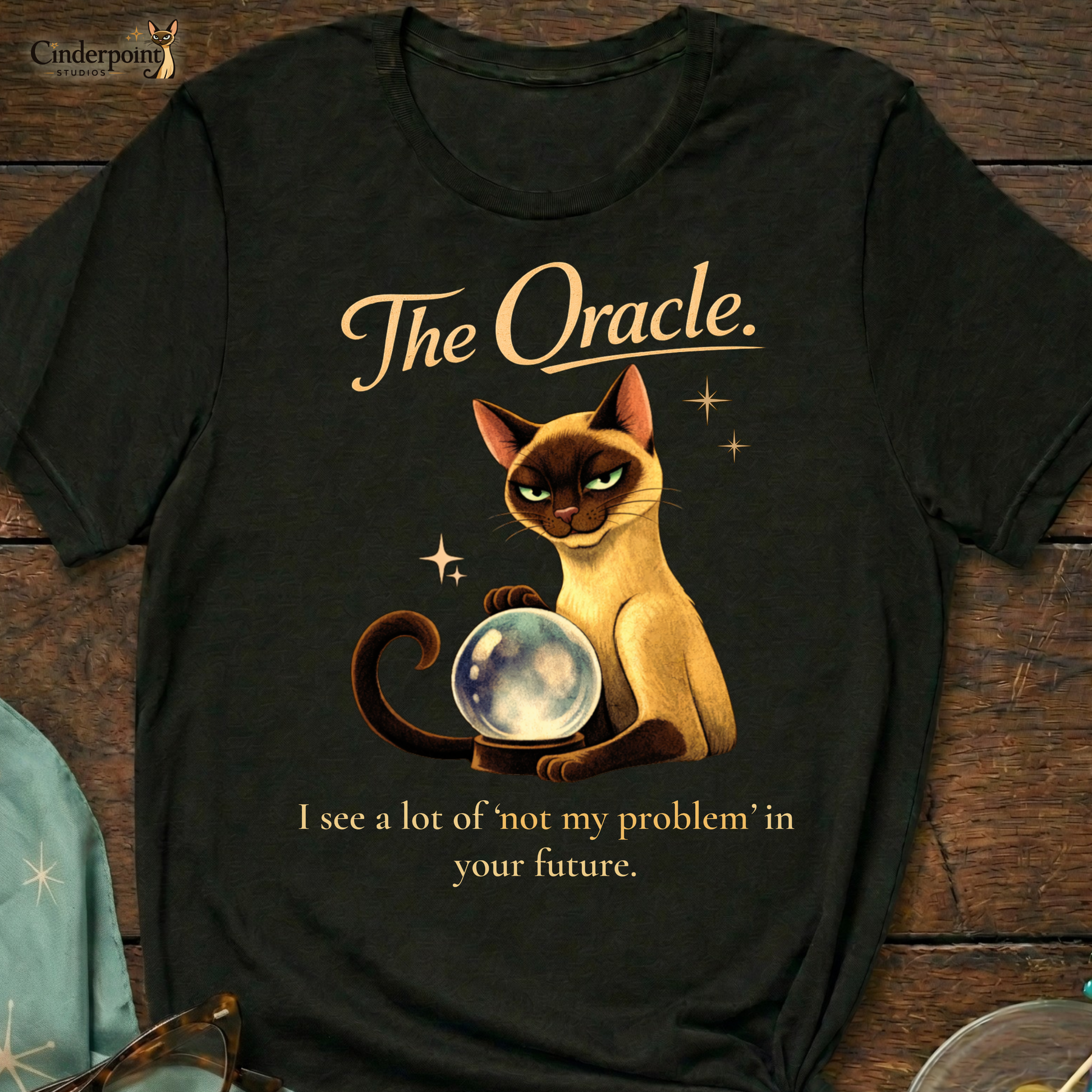 A black Bella+Canvas 3001 t-shirt featuring "The Oracle" design on a dark wood grain table. The design shows a Siamese cat surrounded by atomic stars and the text "The Oracle" and "I see your problem. I also see that it’s not mine." Accessories include vintage cat-eye glasses and a starburst scarf. Cinderpoint Studios logo in the corner.