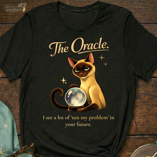 A black Bella+Canvas 3001 t-shirt featuring "The Oracle" design on a dark wood grain table. The design shows a Siamese cat surrounded by atomic stars and the text "The Oracle" and "I see your problem. I also see that it’s not mine." Accessories include vintage cat-eye glasses and a starburst scarf. Cinderpoint Studios logo in the corner.