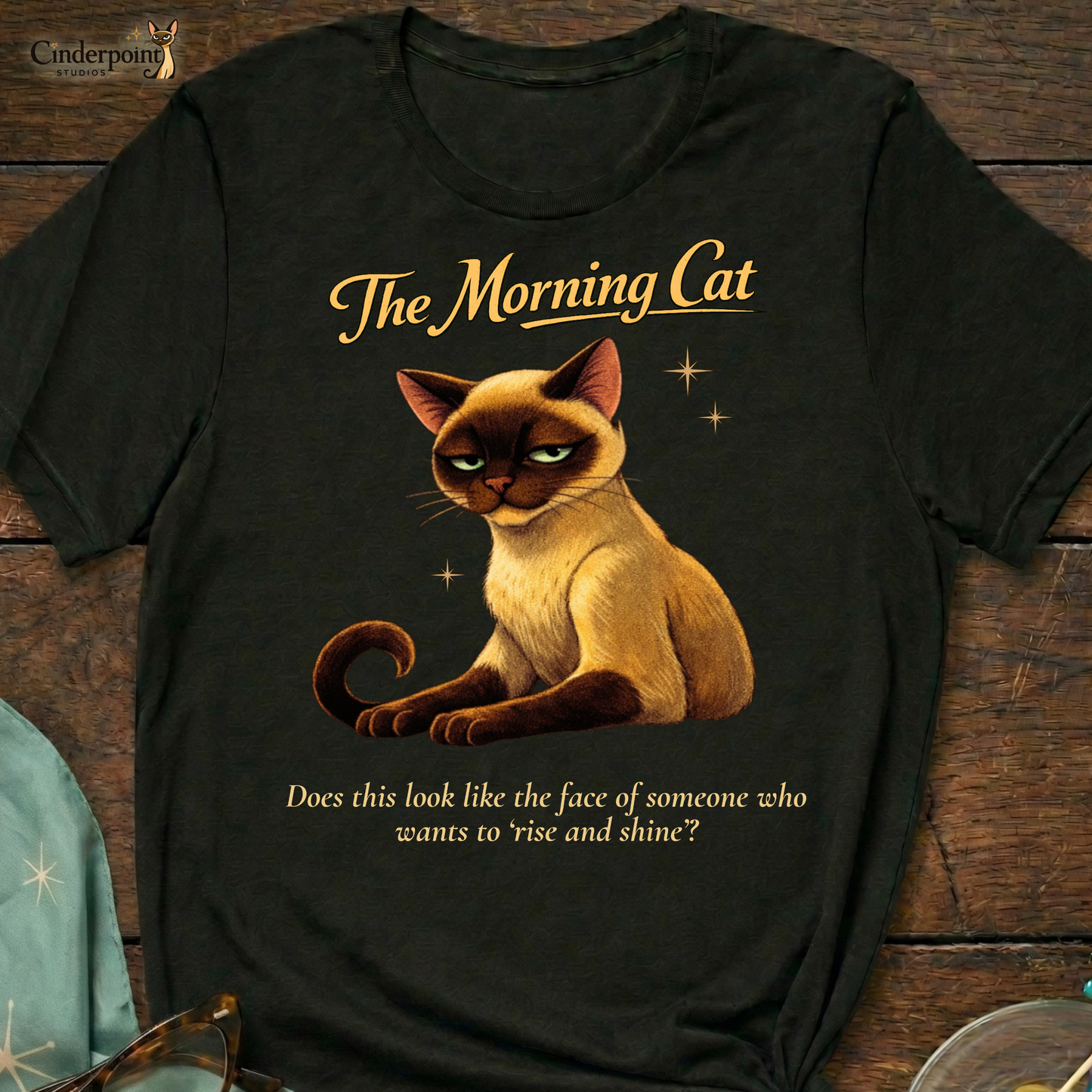 A black Bella+Canvas 3001 t-shirt featuring "The Morning Cat" design on a dark wood grain table. The design shows an exhausted Siamese cat with narrowed eyes and the text "The Morning Cat" and "Does this look like the face of someone who wants to ‘rise and shine’?" Accessories include vintage cat-eye glasses and mid-century modern decor.