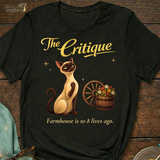 A black Bella+Canvas 3001 t-shirt featuring "The Critique" design. The artwork shows Lyle the Siamese cat recoiling in horror from a rustic wagon wheel and flower barrel. The text reads "The Critique" and "Farmhouse is so 8 lives ago." Placed on a dark wood grain surface with vintage barware and atomic starburst accessories.