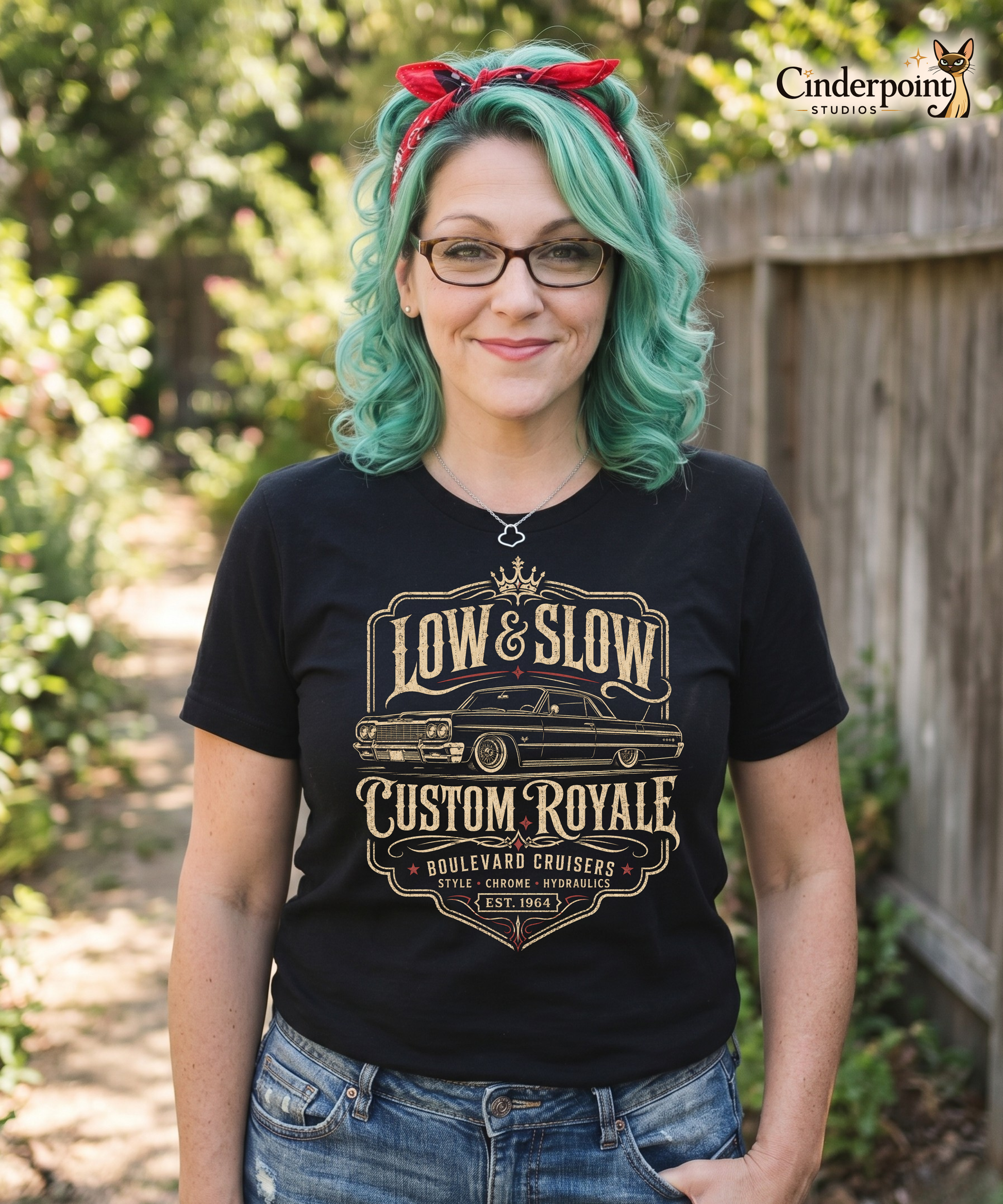 Teal haired woman wearing a black graphic tee featuring original Cinderpoint Studios artwork for Low & Slow Custom Royale, a retro lowrider design with a long custom cruiser inspired by a 1964 Impala, bold vintage lettering, crown detail, elegant framing, and Boulevard Cruisers text.