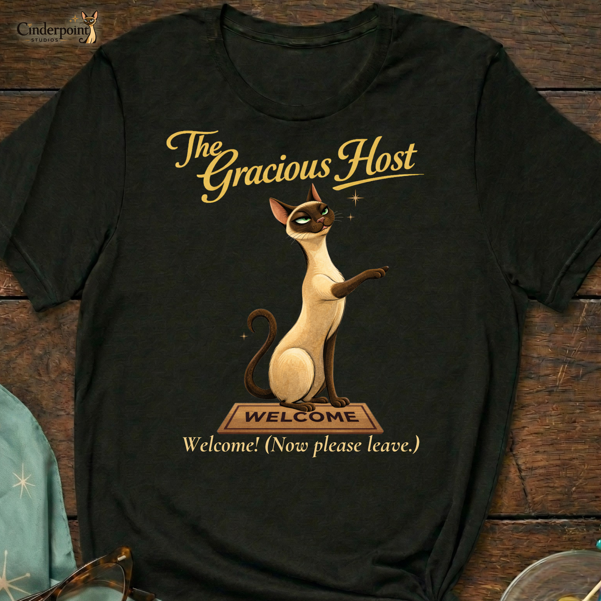A black Bella+Canvas 3001 t-shirt featuring "The Gracious Host" design. The artwork shows Lyle the Siamese cat sitting regally behind a wooden "Welcome" mat with a smug, knowing expression. The text reads "The Gracious Host" and "Welcome! (Now please leave.)" placed on a dark wood grain surface with vintage accessories.