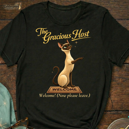 A black Bella+Canvas 3001 t-shirt featuring "The Gracious Host" design. The artwork shows Lyle the Siamese cat sitting regally behind a wooden "Welcome" mat with a smug, knowing expression. The text reads "The Gracious Host" and "Welcome! (Now please leave.)" placed on a dark wood grain surface with vintage accessories.