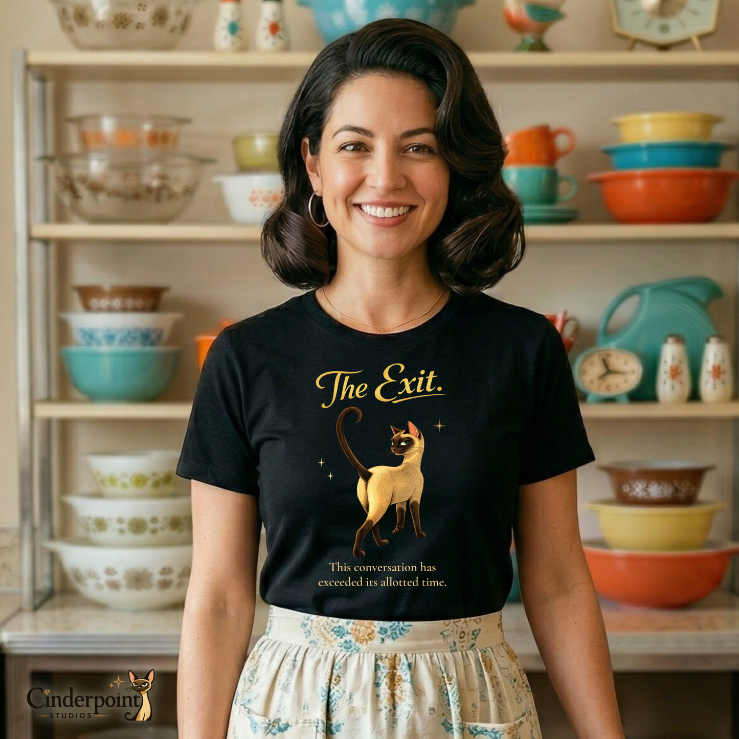 A smiling woman in a retro 1950s kitchen wearing "The Exit" t-shirt and a patterned half-apron. She stands in front of a hutch filled with colorful Pyrex bowls.