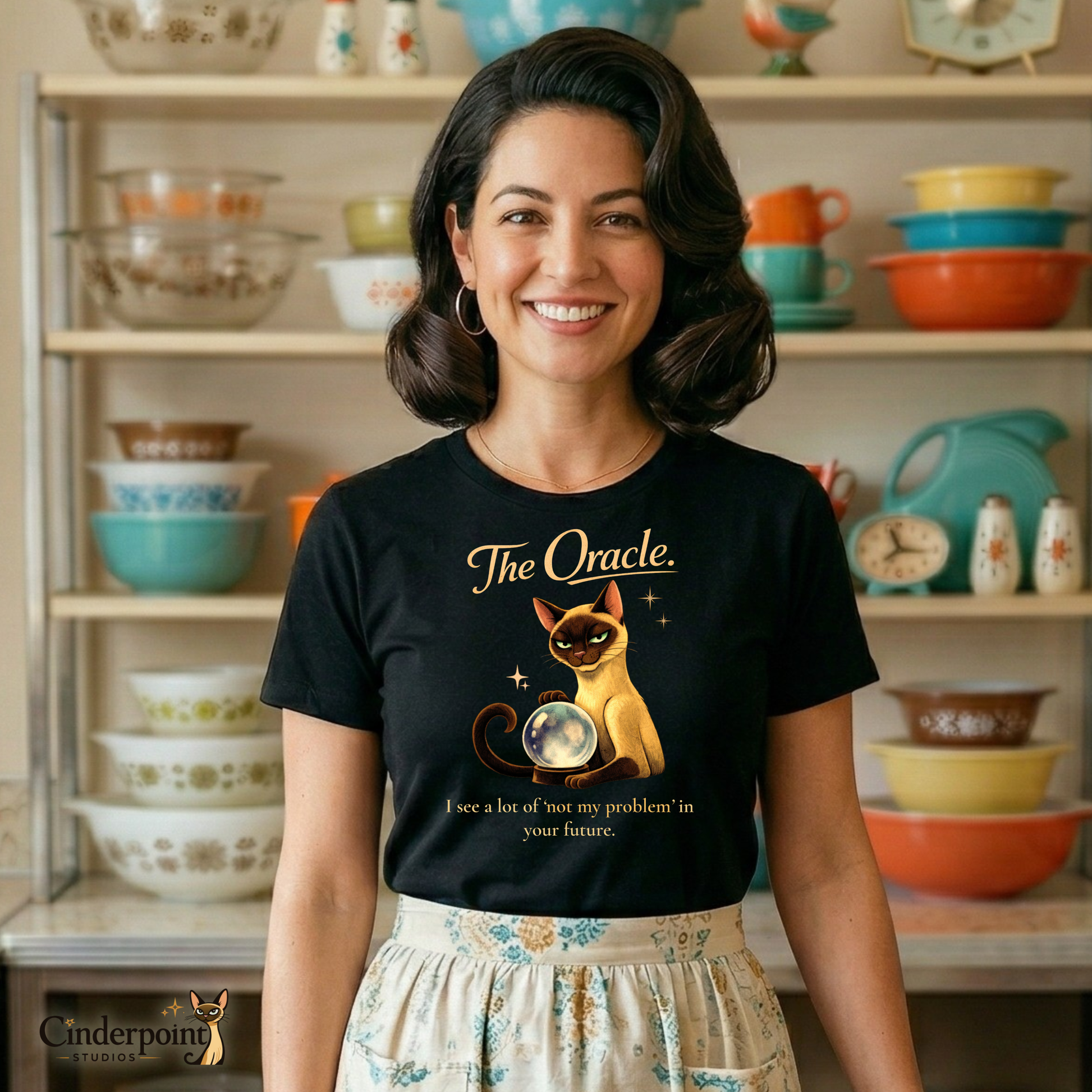 A smiling woman in a retro 1950s kitchen wearing "The Oracle" t-shirt and a patterned half-apron. She stands in front of a hutch filled with colorful Pyrex bowls, blending "Better Living" nostalgia with Lyle's mystical sass.