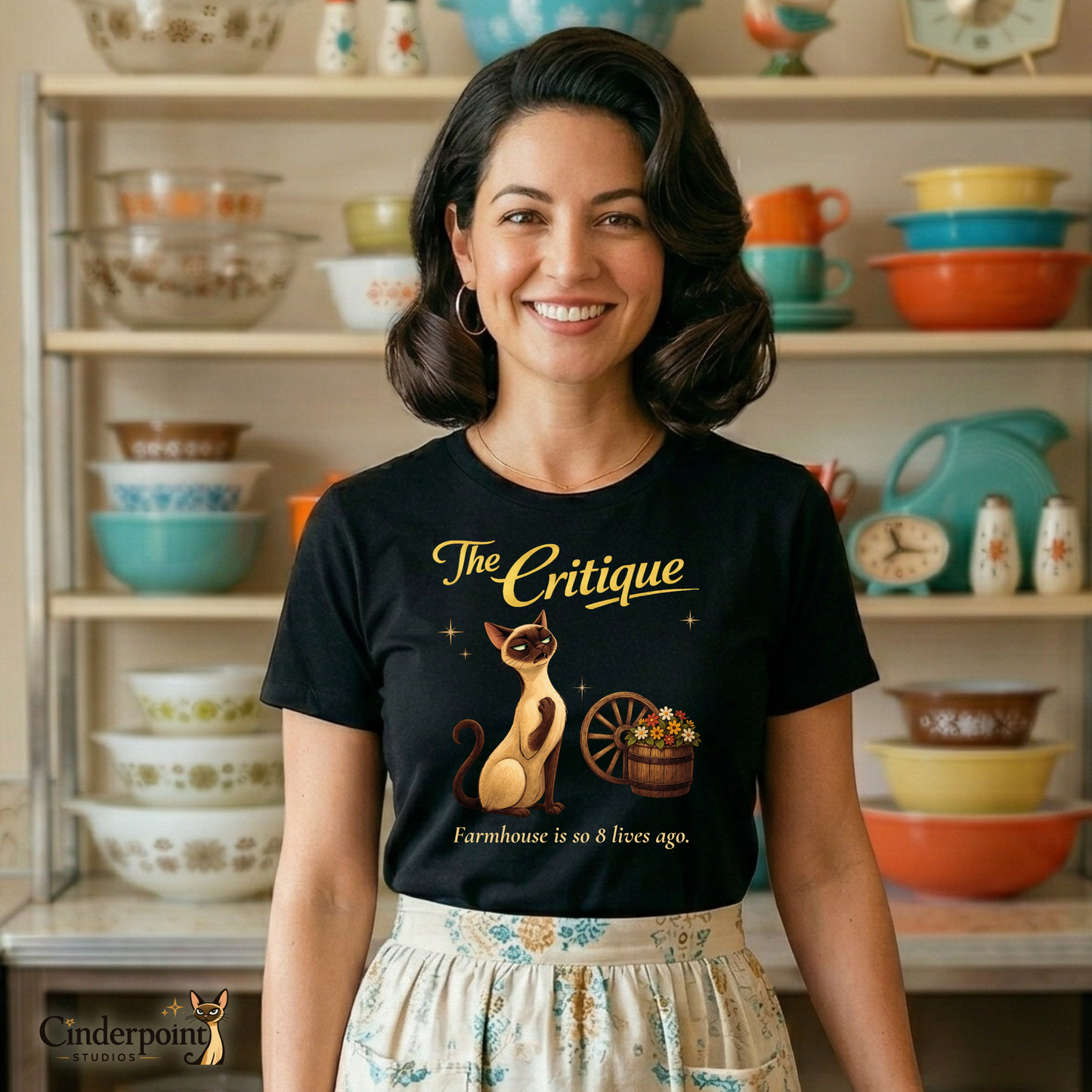 A smiling woman in a retro 1950s kitchen wearing "The Critique" t-shirt and a patterned half-apron. She stands in front of her pristine Pyrex collection, highlighting the "anti-farmhouse" vibe of the design.