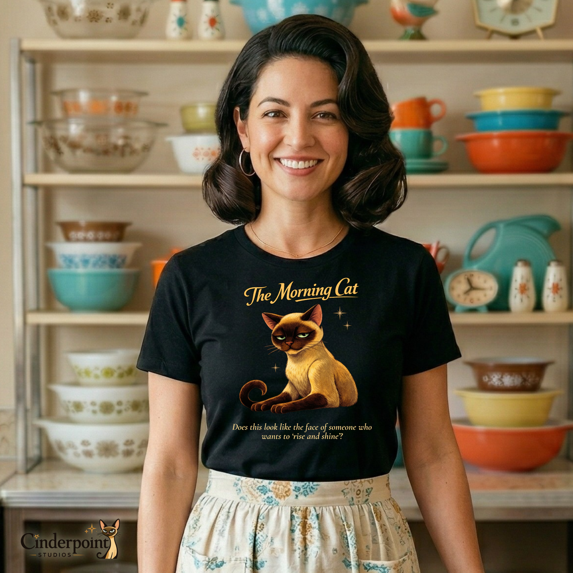 A woman in a retro 1950s kitchen wearing "The Morning Cat" t-shirt and a patterned half-apron. She stands in front of her Pyrex collection with an expression that says she is not ready to cook breakfast.
