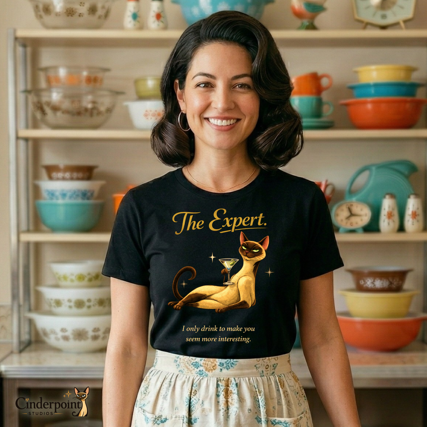 A smiling woman in a retro 1950s kitchen wearing "The Expert" t-shirt and a patterned half-apron. She stands in front of a hutch filled with colorful Pyrex bowls, capturing a "Better Living" nostalgic vibe.