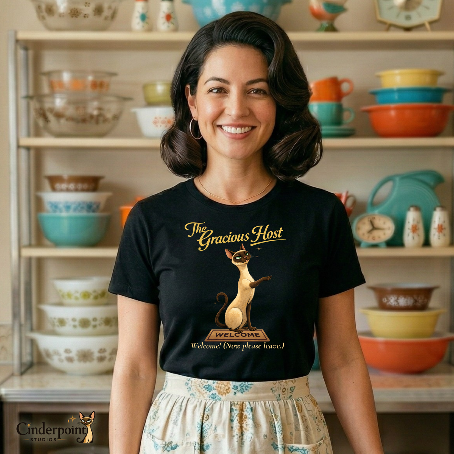 A smiling woman in a retro 1950s kitchen wearing "The Gracious Host" t-shirt and a patterned half-apron. She stands in front of a hutch filled with colorful Pyrex, highlighting the "Better Living" nostalgia of the series.