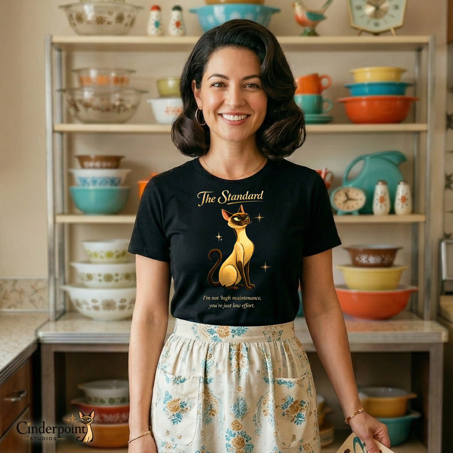 A smiling woman with a vintage bob hairstyle, standing in a detailed 1950s atomic-style kitchen. She is wearing a black Bella+Canvas 3001 t-shirt featuring "The Standard" Lyle the cat design, paired with a patterned vintage half-apron. She is holding a small patterned plate. Behind her, a metal hutch displays a collection of colorful stacked Pyrex bowls and other retro kitchenware.