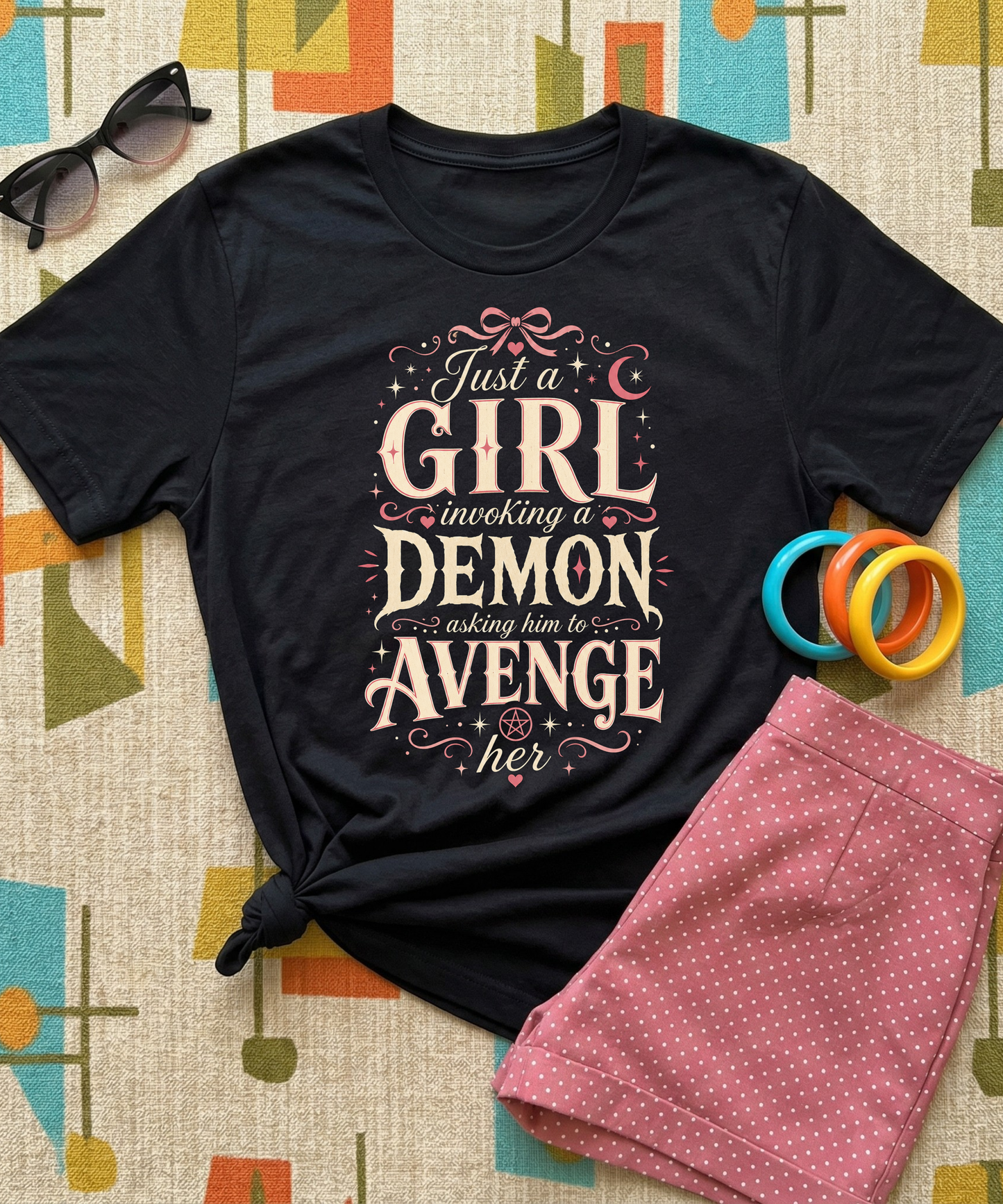 Black graphic tee featuring original Cinderpoint Studios artwork for Just a Girl Invoking a Demon, a feminine occult humor design with bold vintage lettering, pink bow accents, crescent moon, stars, hearts, and a small pentagram.