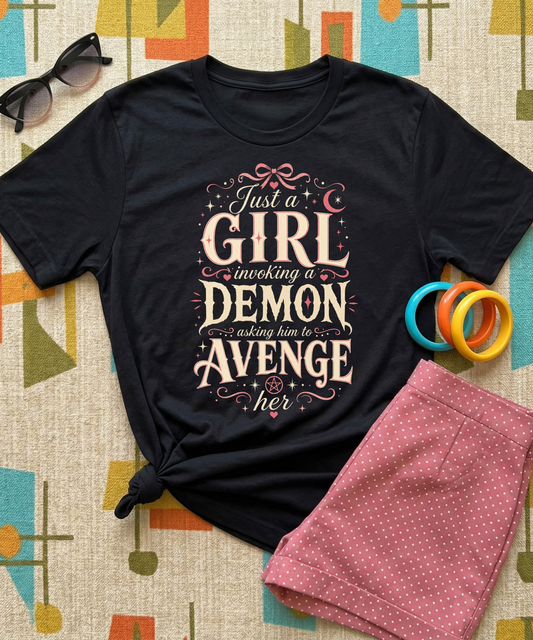Black graphic tee featuring original Cinderpoint Studios artwork for Just a Girl Invoking a Demon, a feminine occult humor design with bold vintage lettering, pink bow accents, crescent moon, stars, hearts, and a small pentagram.